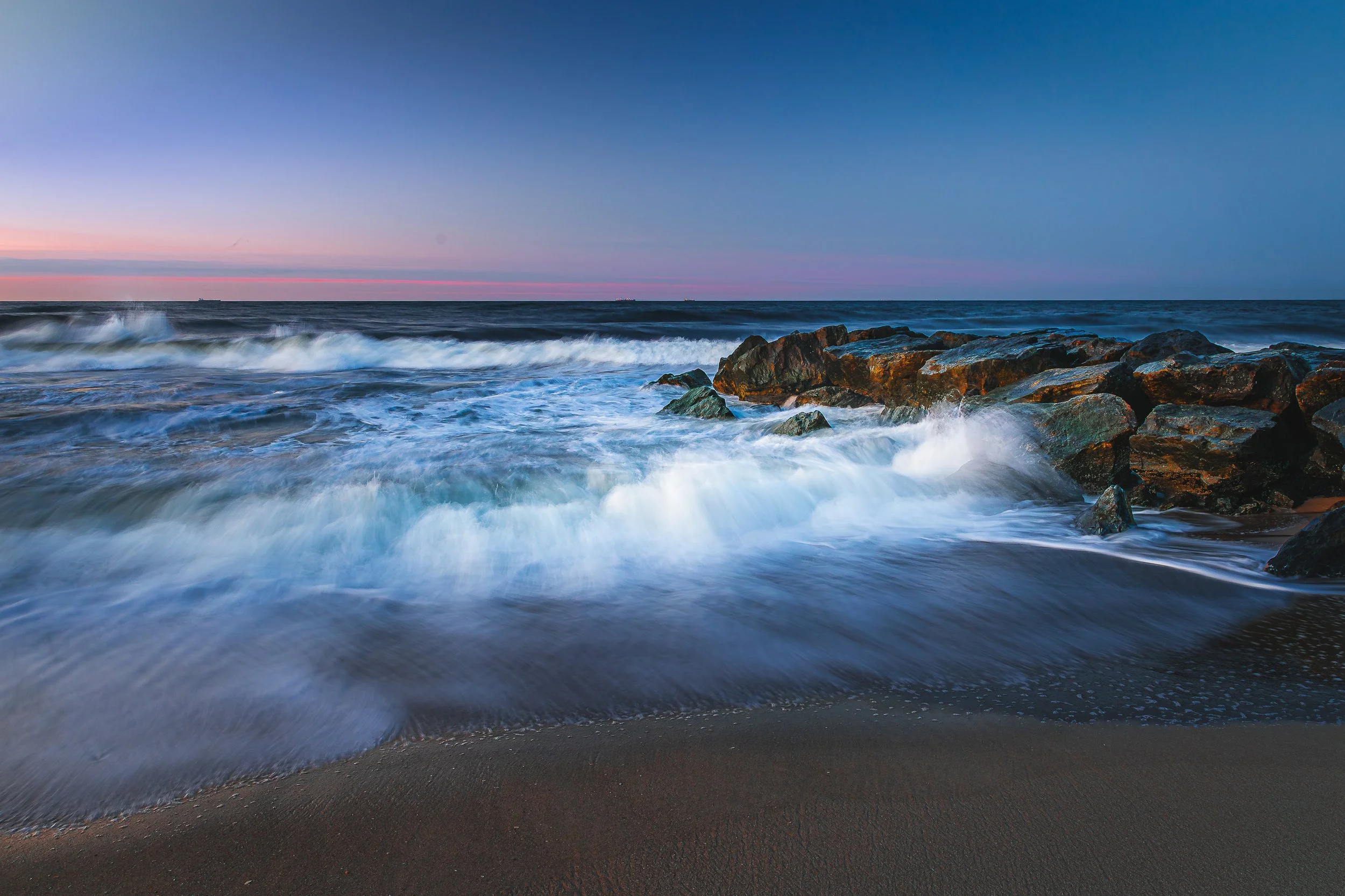 Photo of ocean waves crashing against rocks on a sandy beach during sunset or sunrise, with a clear sky and a few ships on the horizon.