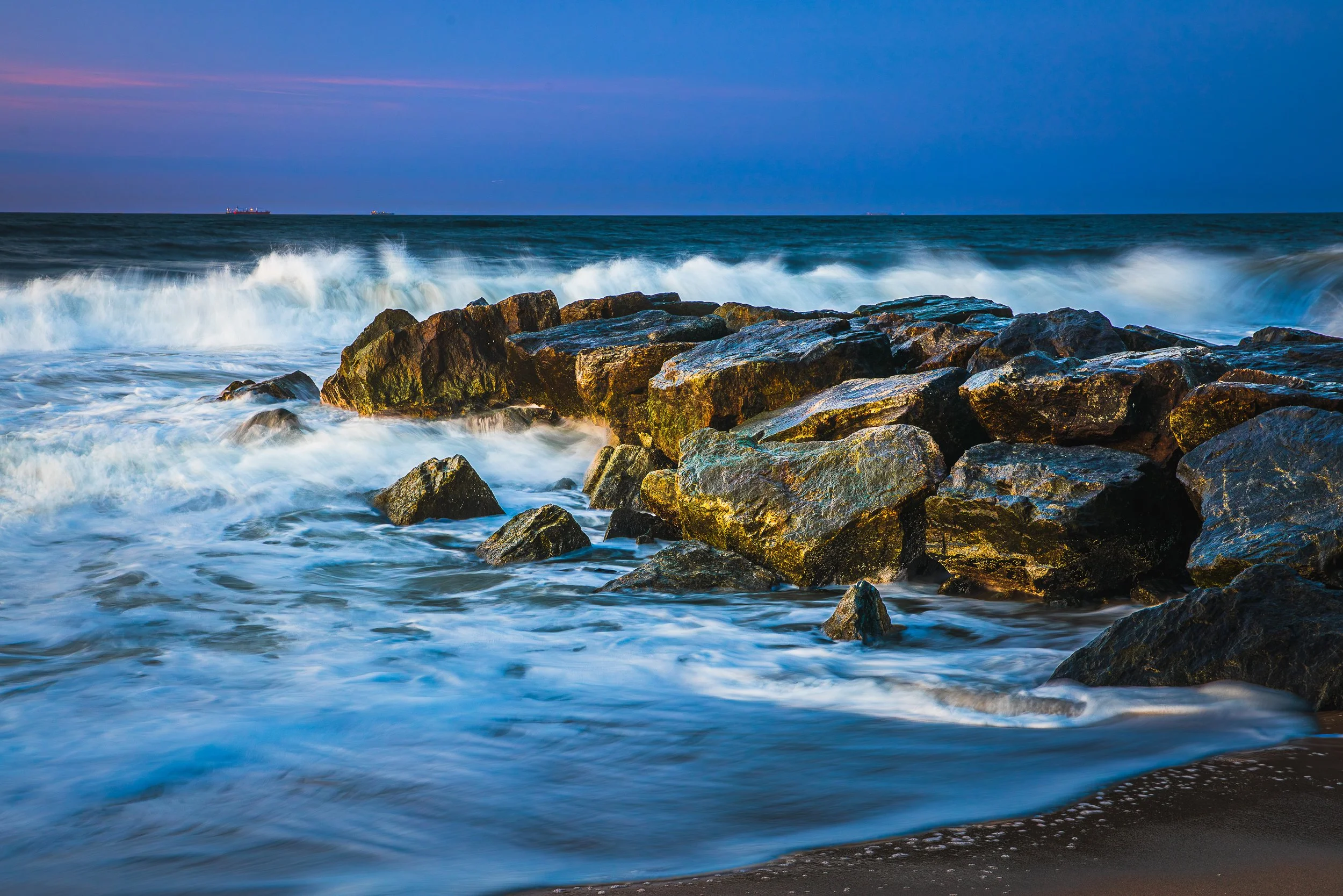 Ocean waves crashing against rocks on a beach at sunset or sunrise.