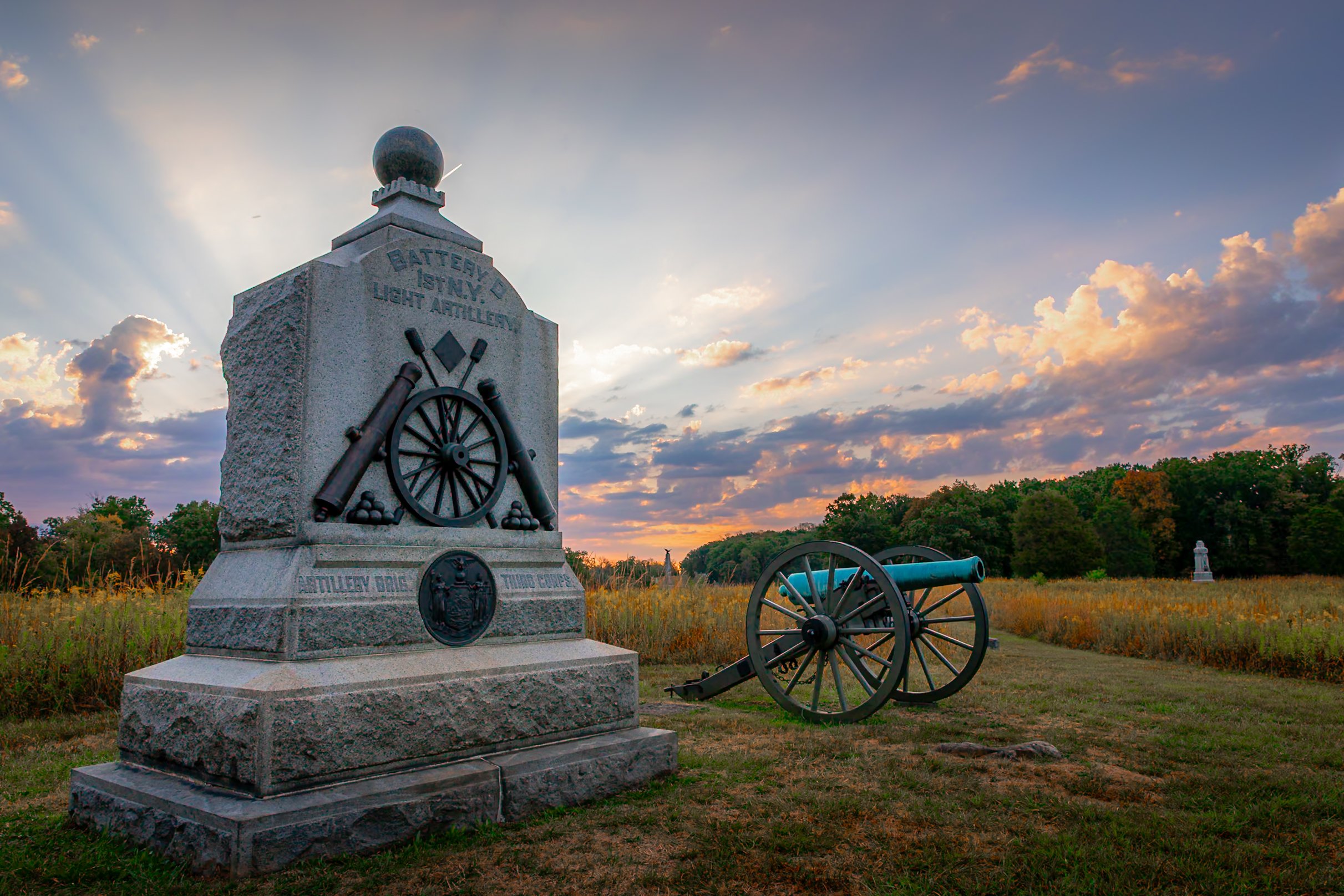 A stone monument with cannons and artillery symbols at sunrise in Gettysburg National Battlefield , marked 'Battery' and 'Light Artillery,' with a cannon nearby and a field with trees in the background.