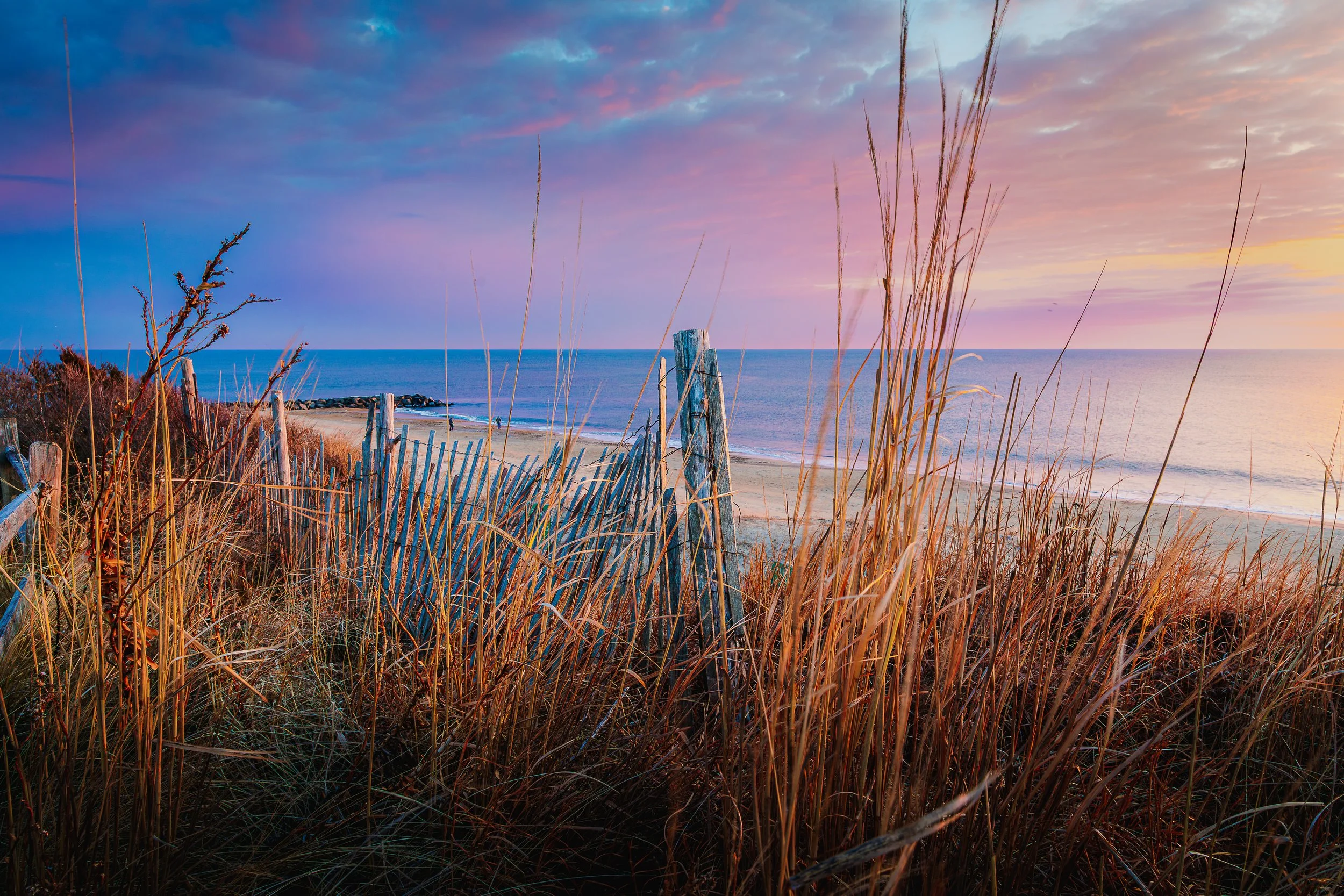 Beach scene during sunset with tall grasses in the foreground, a weathered wooden fence, calm ocean waves, and colorful sky with clouds.