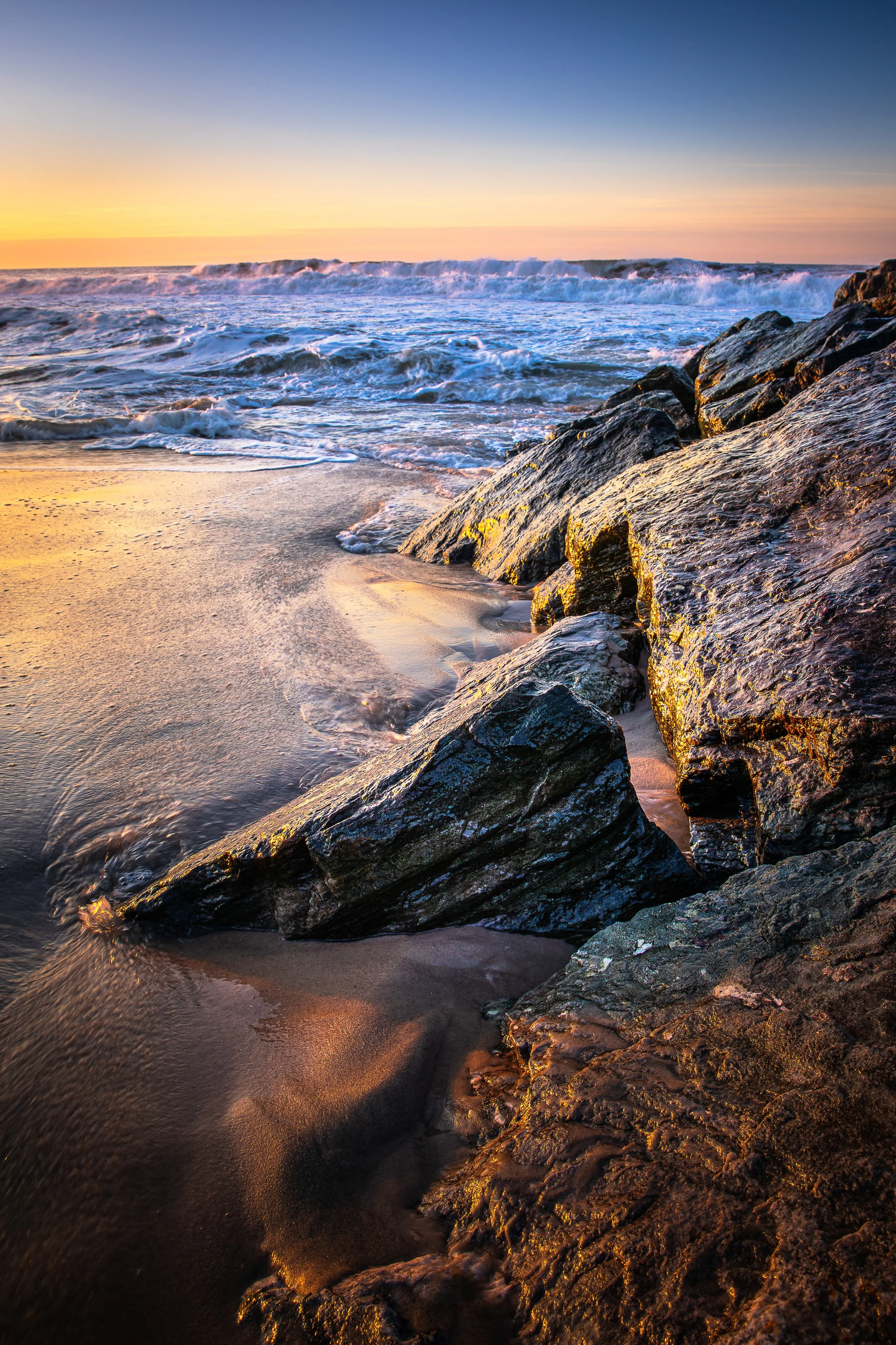 Sunset over a rocky beach with waves crashing and wet sand glowing in the light.