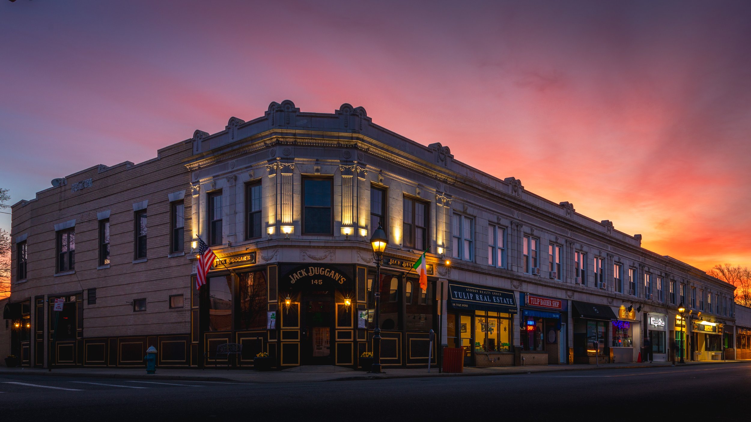 Historic building on a street corner In Floral Park NY at sunrise, with illuminated windows and signs for local shops and restaurants, including Jack Duggans, Tulip Real Estate, and Tulip Barber Shop.