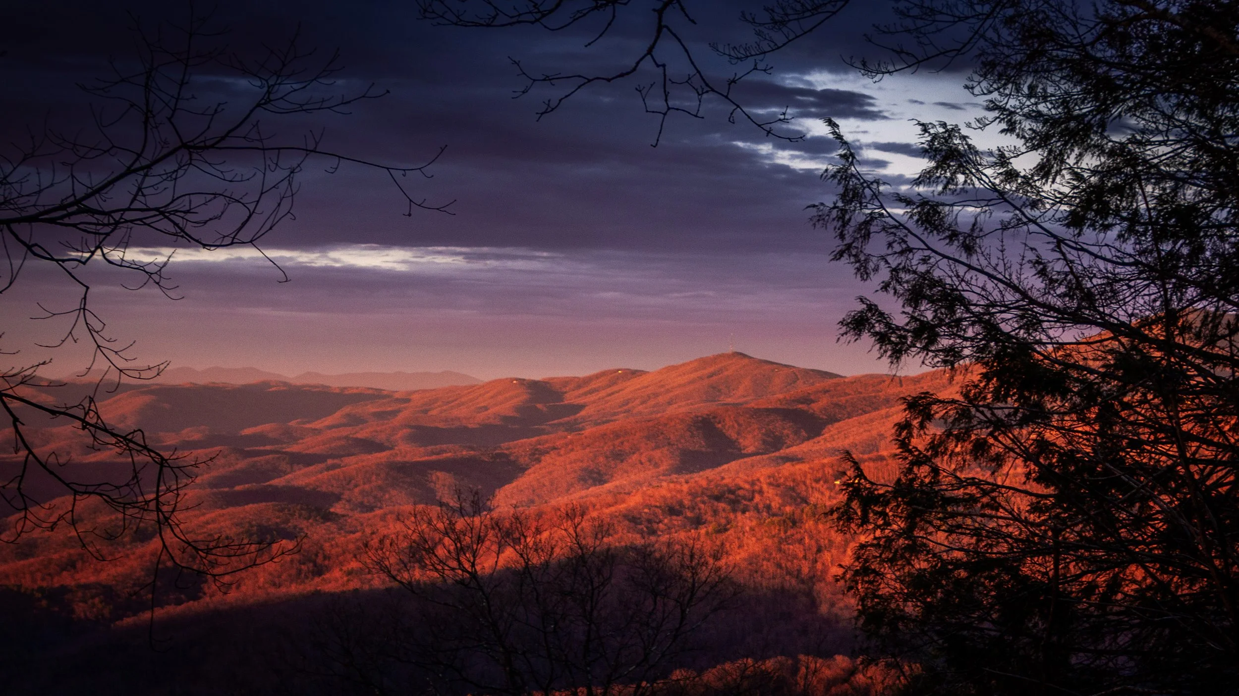 Sunrise over the Blue Ridge mountain range in Blowing Roak NC with orange and purple hues, silhouetted tree branches in foreground, cloudy sky.