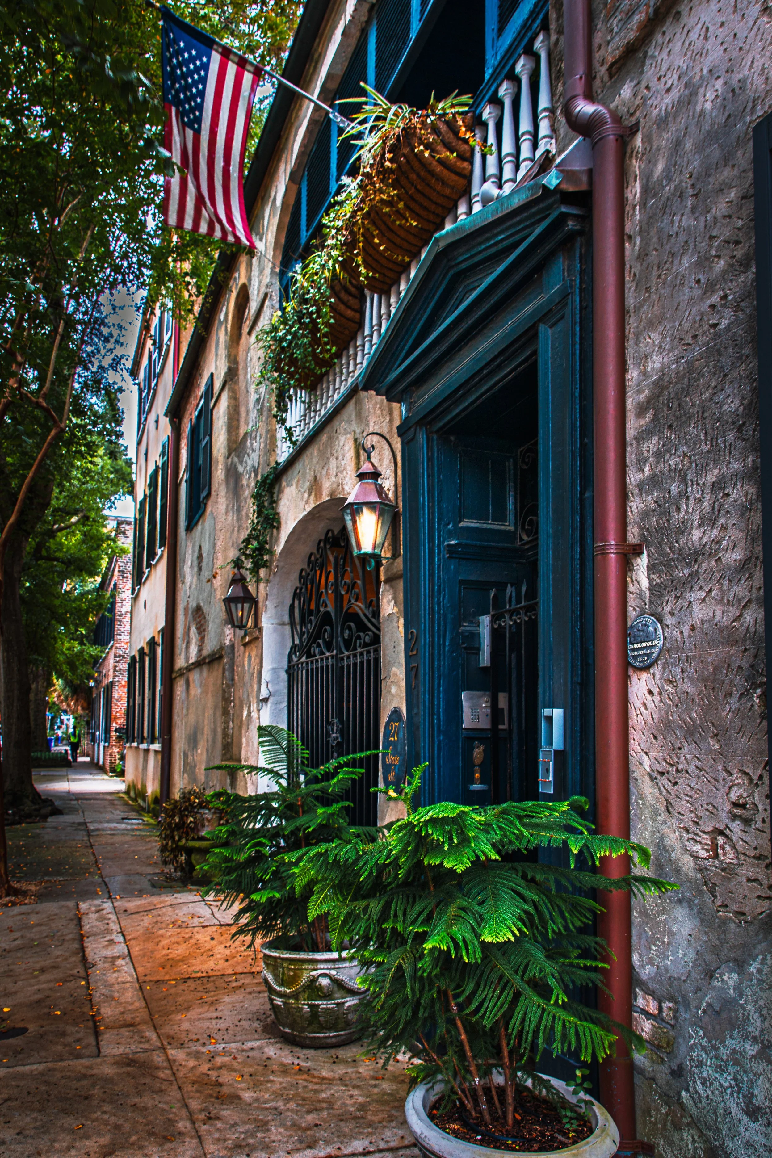 A narrow sidewalk in Charleston SC lined with potted plants and an old building with a dark blue door, iron gate, and a flag of the United States hanging from a balcony overhead.