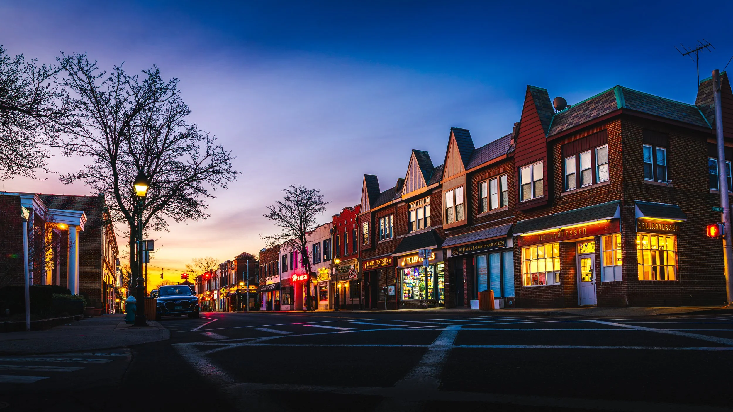 A colorful street scene at sunrise In Floral Park NY with brick buildings, illuminated storefronts, leafless trees, a parked car, and traffic lights.