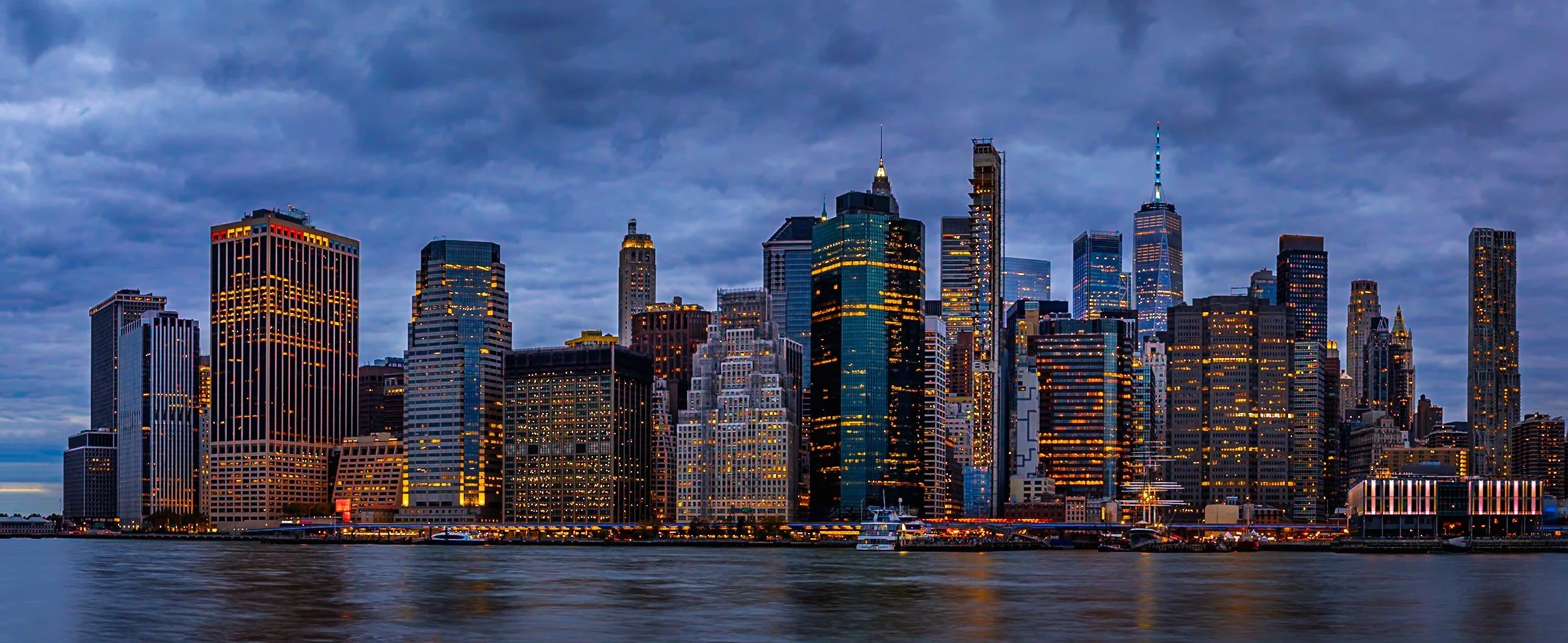 Nighttime view of a city skyline with tall skyscrapers and buildings illuminated, seen across a body of water.