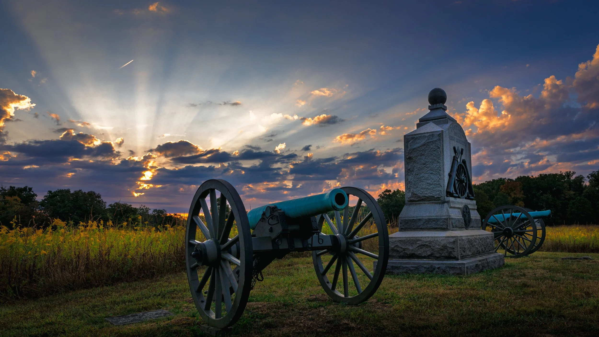 Cannons and a monument at sunset, with a field and trees in the background.