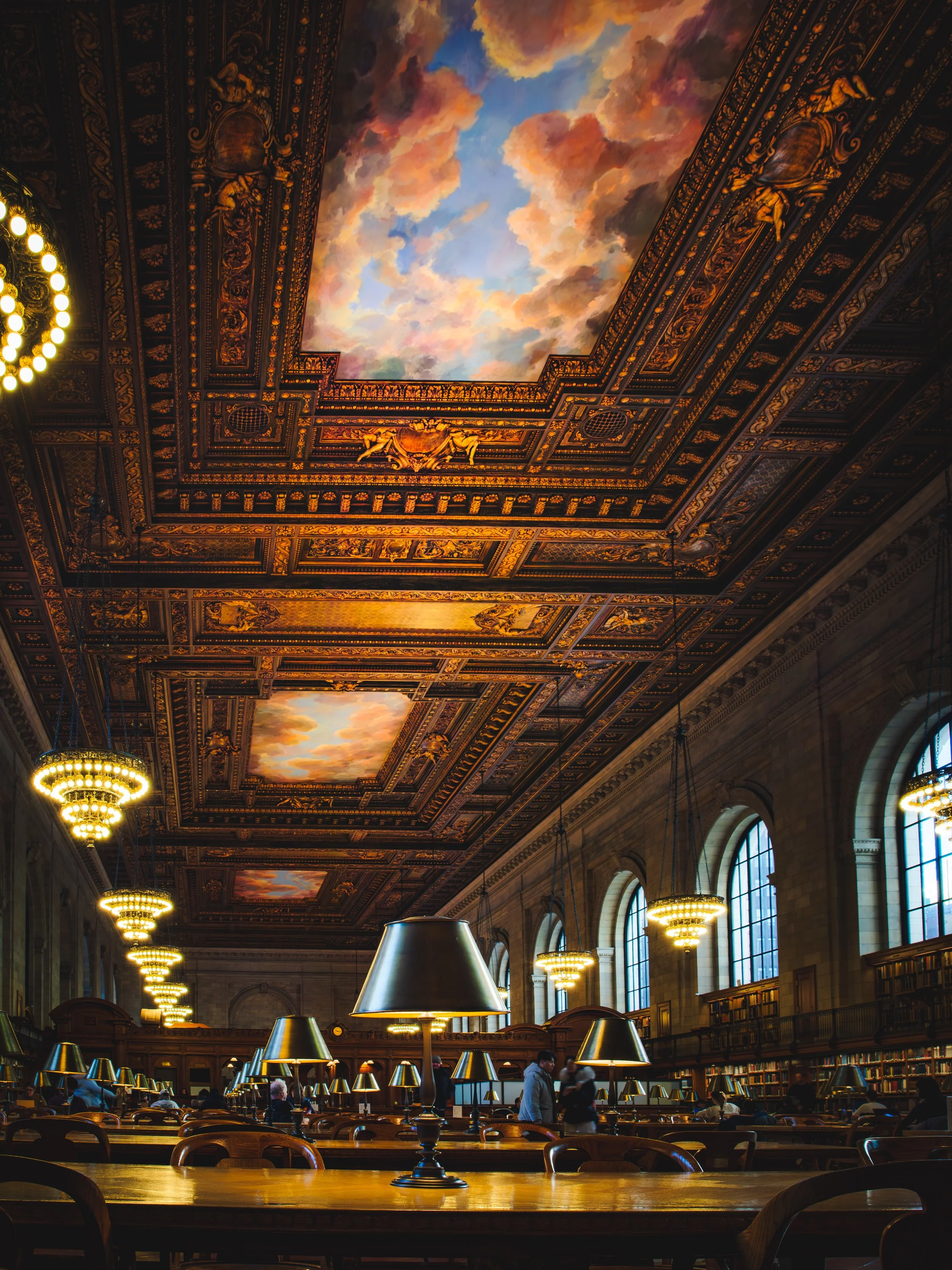 Inside the NY Public Library Rose Reading Room with vaulted ceilings featuring painted clouds, large arched windows, and rows of tables with lamps. Several people are seated and walking inside.
