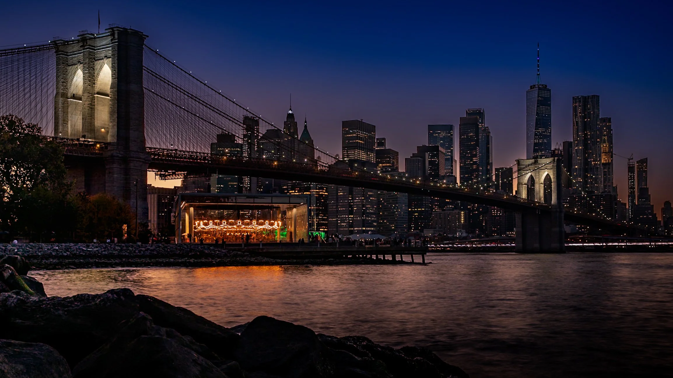 Nighttime view of the Brooklyn Bridge and Manhattan skyline in New York City, with illuminated buildings and a carousel in the foreground.