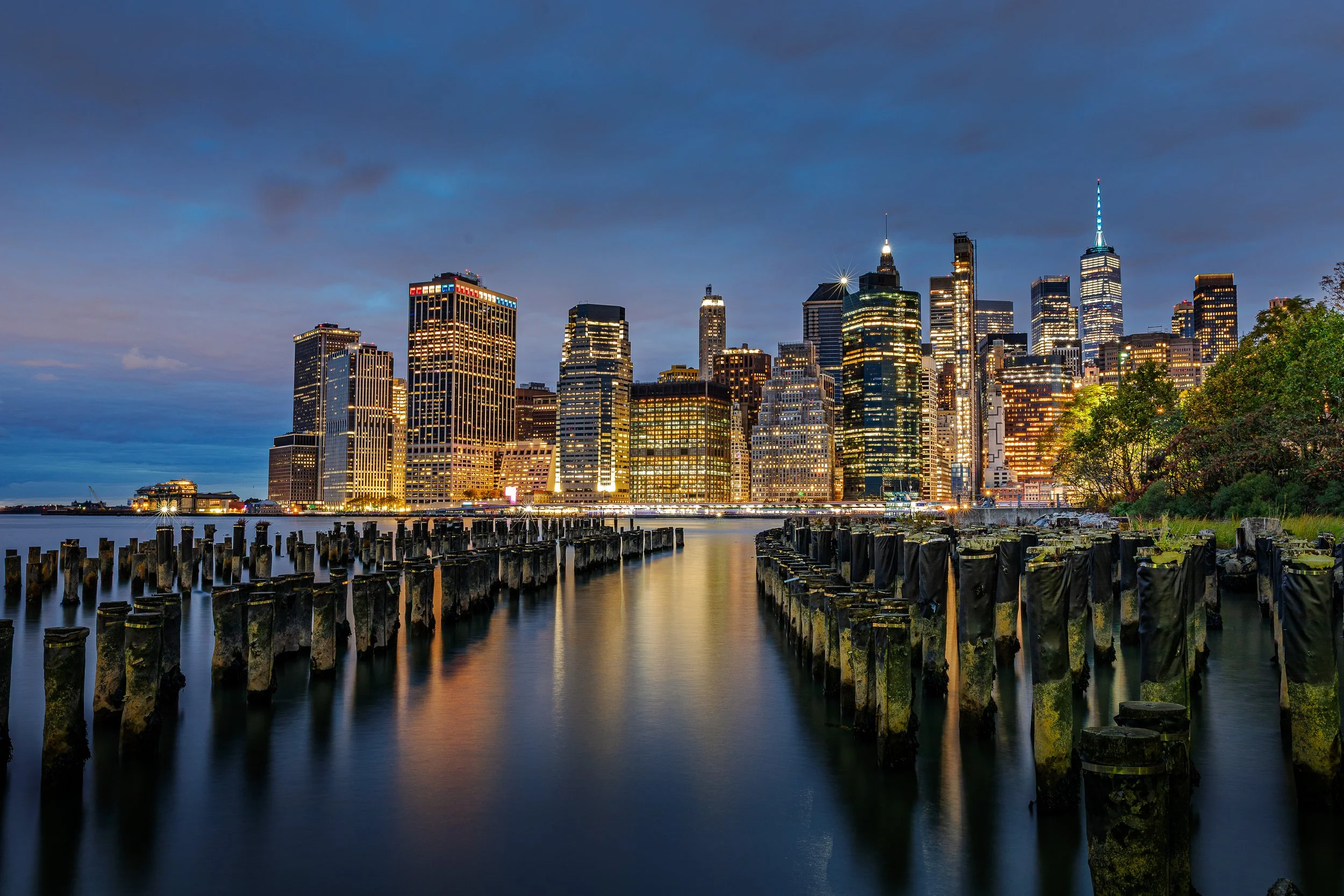 Night view of the New York City skyline with tall illuminated skyscrapers reflecting on the water, with old wooden pilings in the foreground.