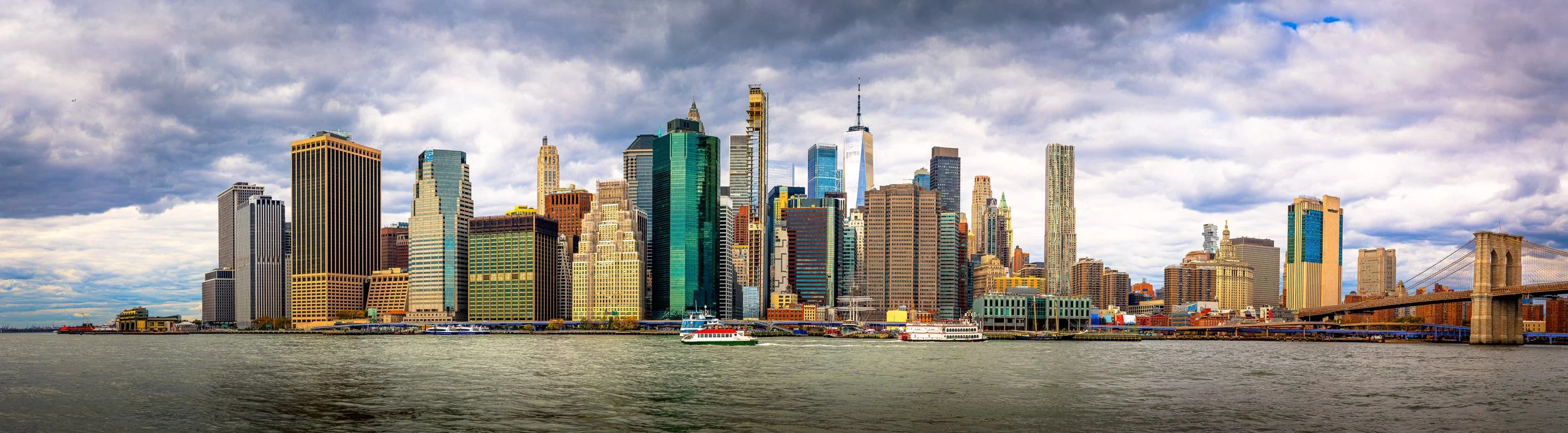 Skyline of Manhattan, New York City, with skyscrapers along the riverfront and a bridge on the right, over water and cloudy sky.
