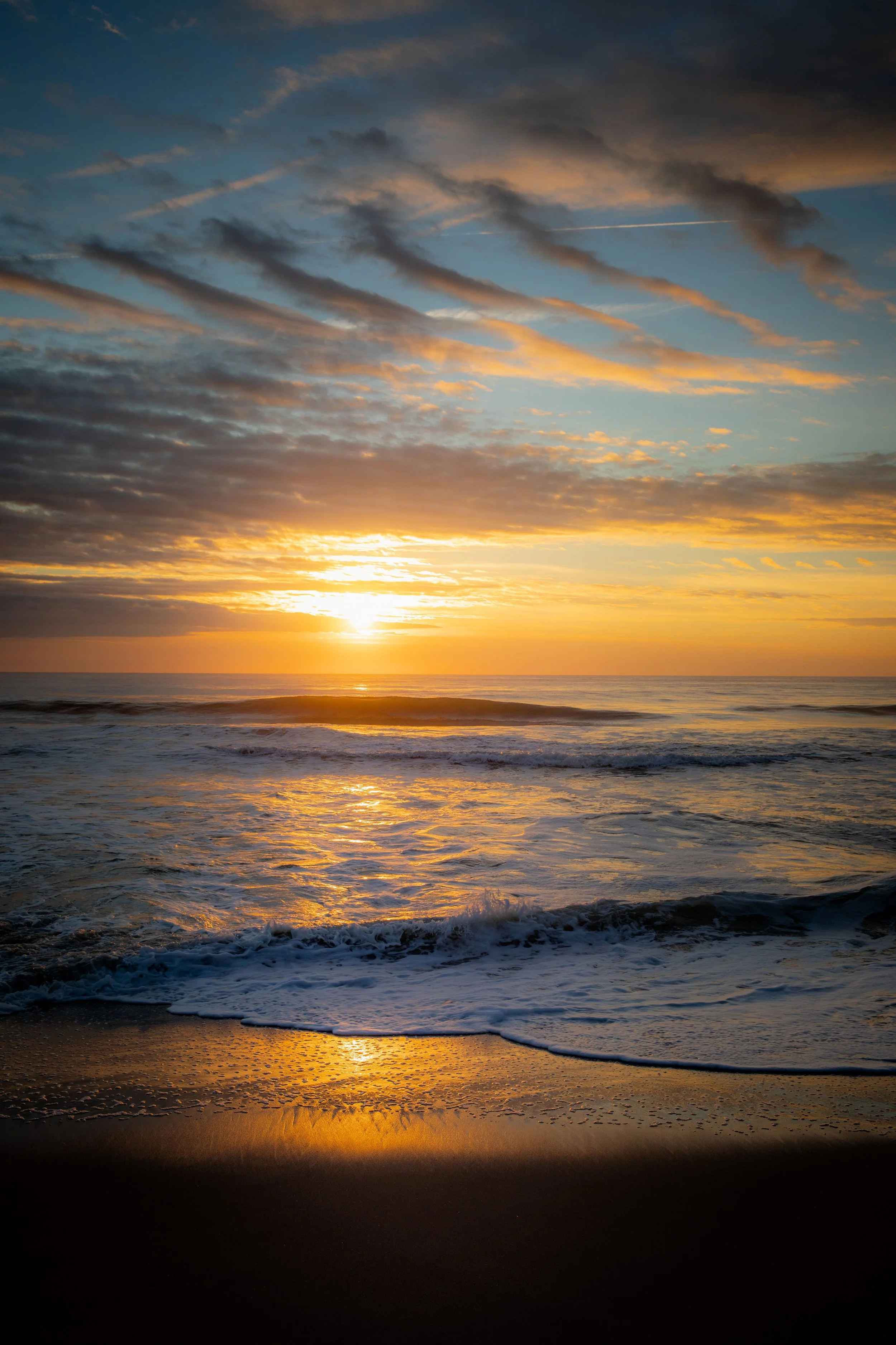 Sunset over the ocean with a cloudy sky and waves crashing onto the sandy shore.