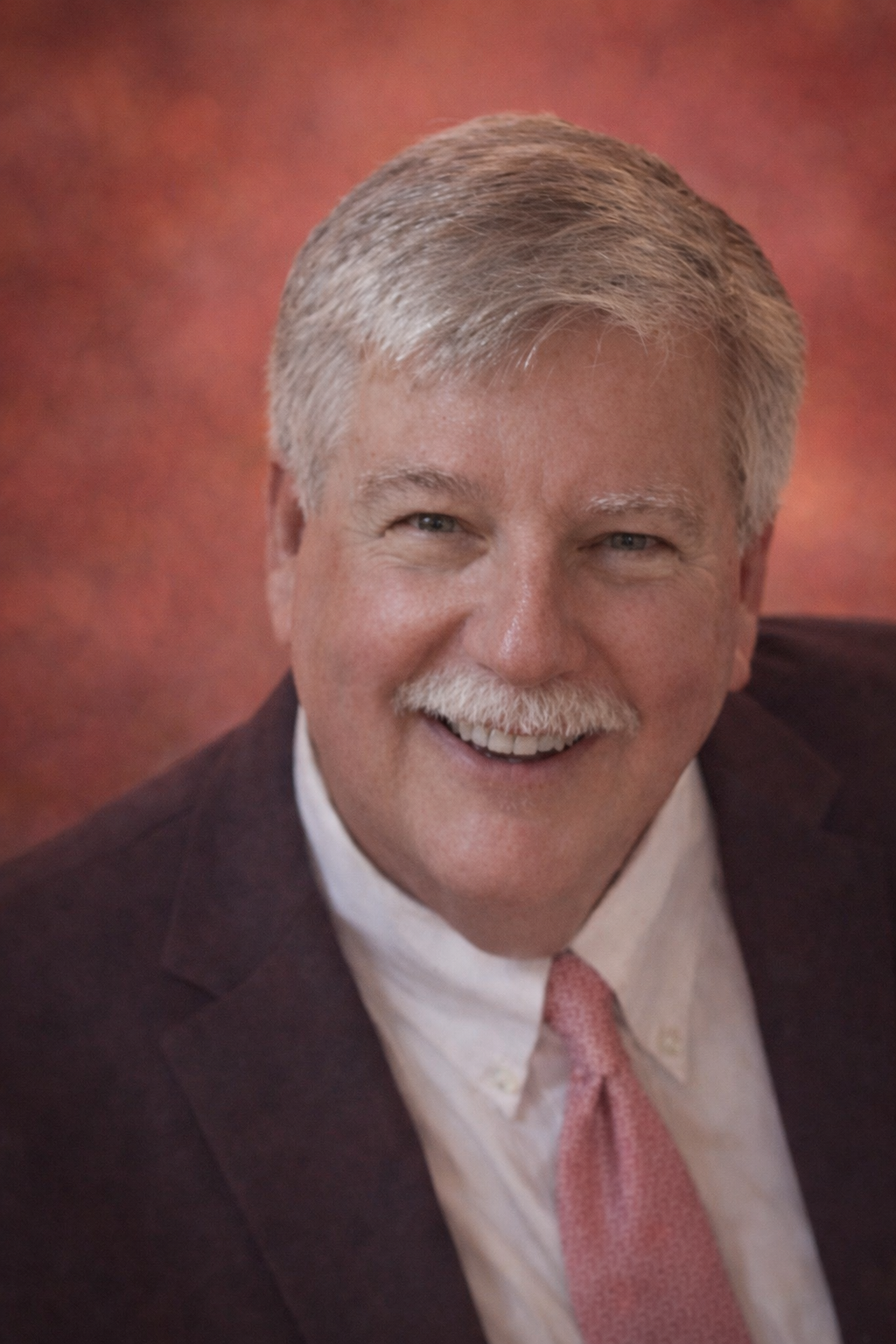 Headshot of a smiling middle-aged man with gray hair, mustache, wearing a dark suit, white shirt, and pink tie.