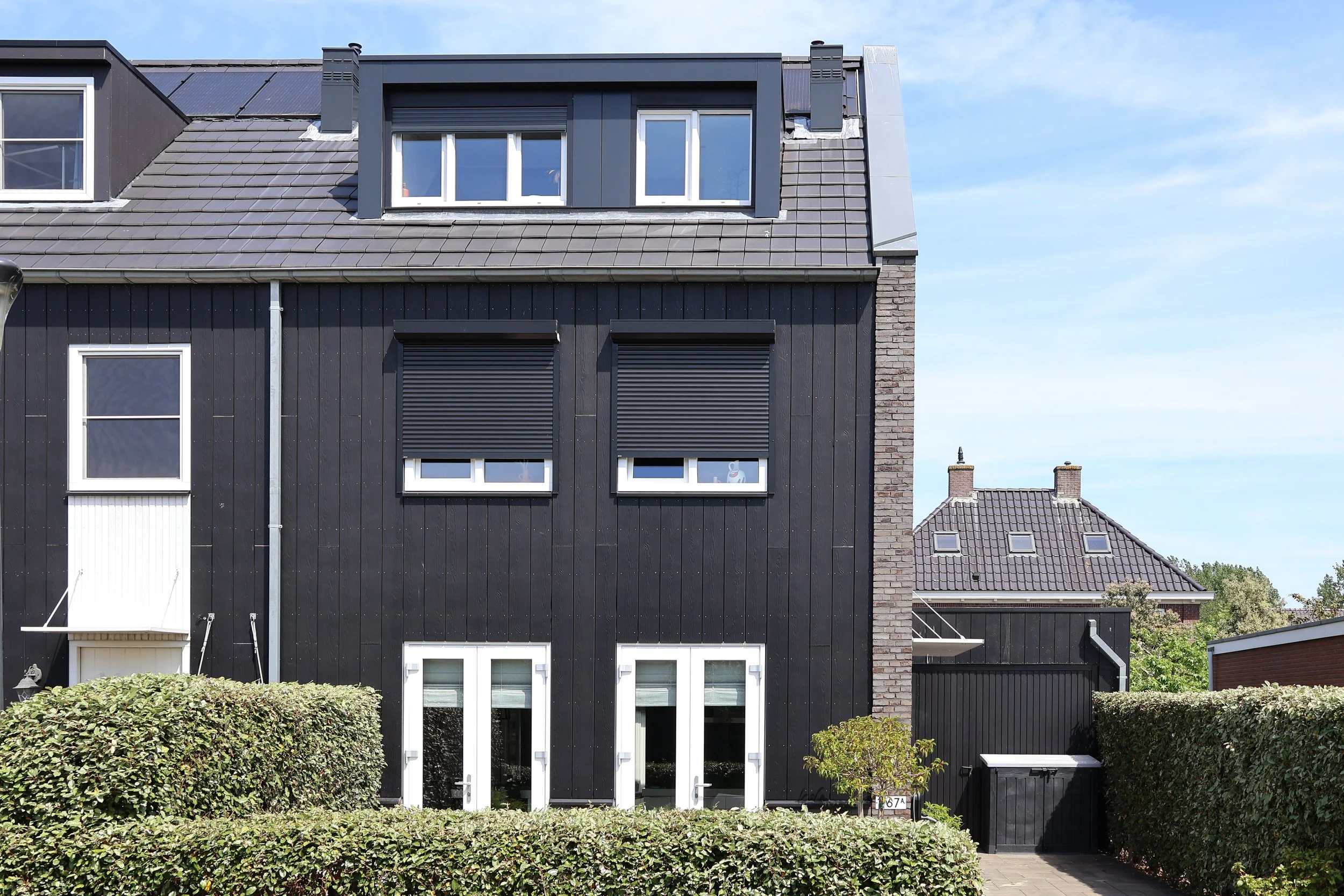 Uitzicht op een modern zwart houten huis met meerdere verdiepingen en grote ramen, begroeid met een groene heg voor de voordeur, onder een blauwe lucht.