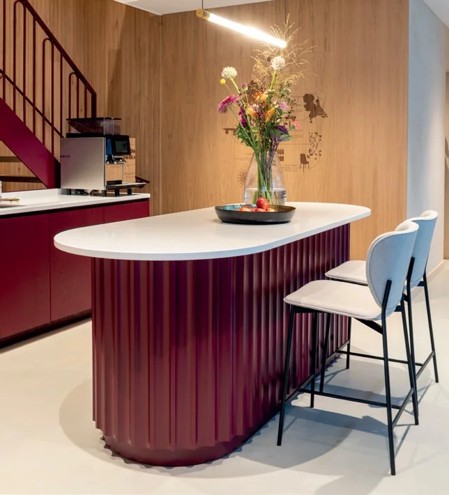 Een modern keukenhoek met een afgeronde bar met een witte bovenkant en een donkerrode ribbenstructuur, twee witte barkrukken, een glazen vaas met bloemen en fruit, en een houten wand op de achtergrond.