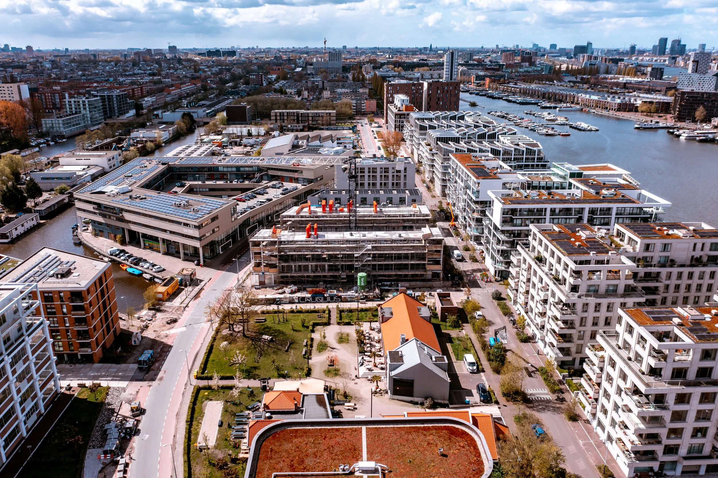 Uitzicht over een stadsgebied met moderne gebouwen, een kanaal met boten en hoogbouw op de achtergrond, onder een bewolkte hemel.