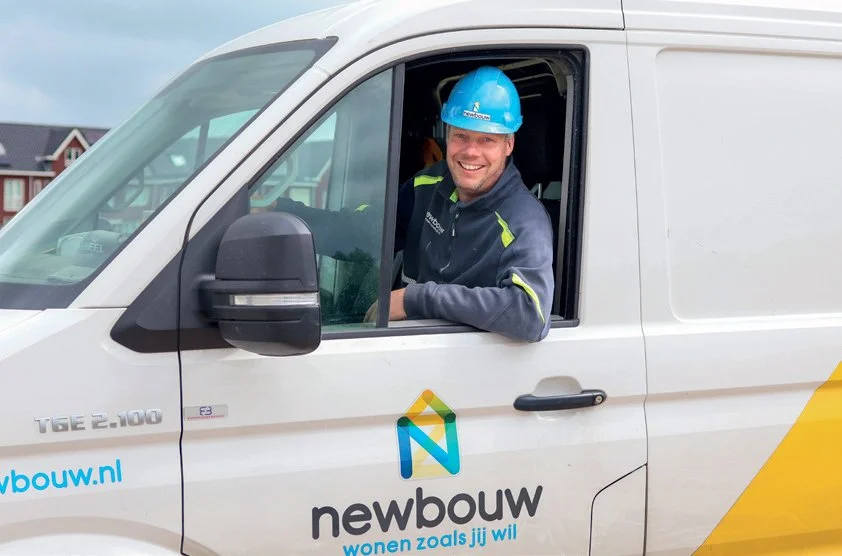 Man in een bouwhelmpje in de bestuurdersstoel van een wit bouwbedrijfvoertuig met logo van newbouw en een glimlach.