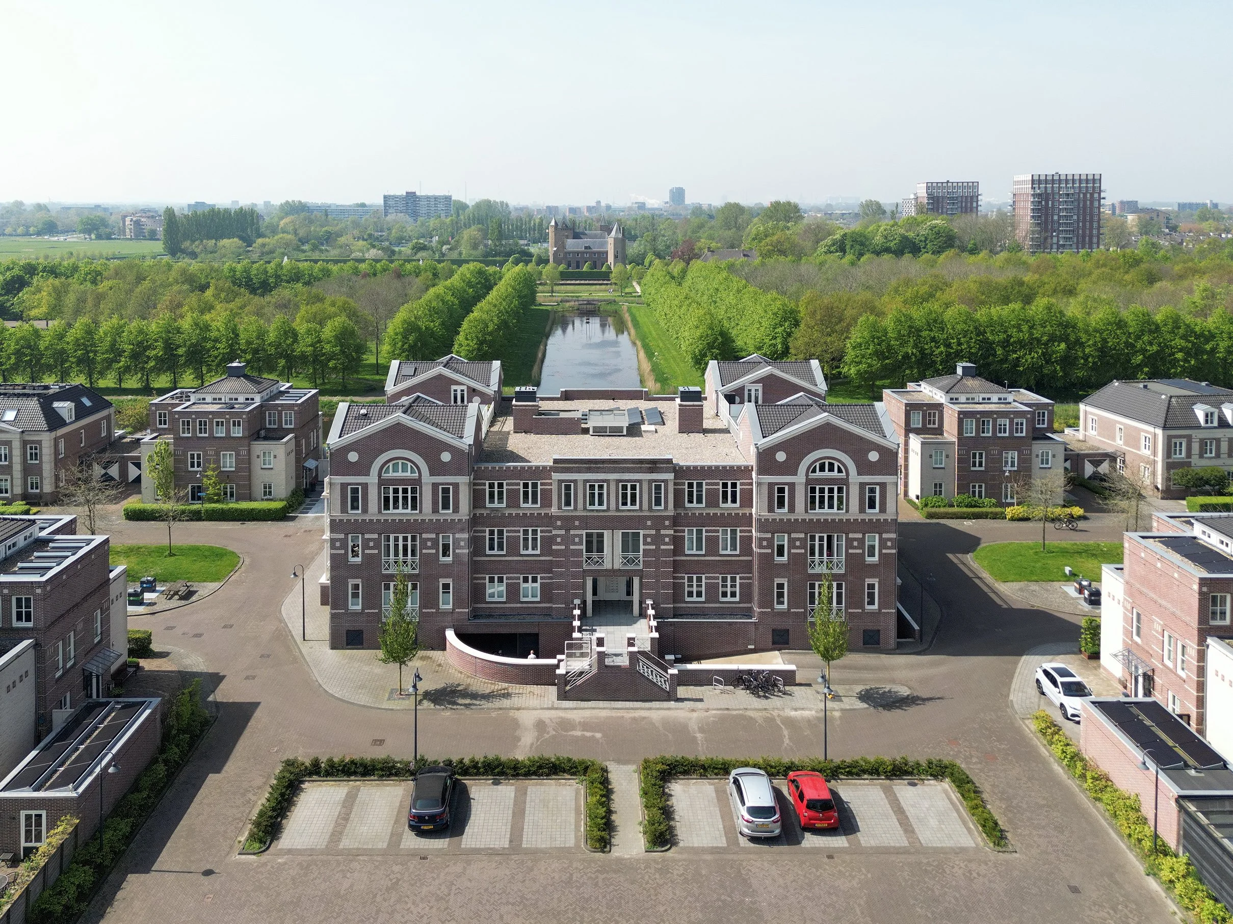 Een overzicht van een stedelijk landschap met klassieke en moderne gebouwen, een smalle slotgracht met brug, omgeven door groen en parkeergelegenheid op de voorgrond.