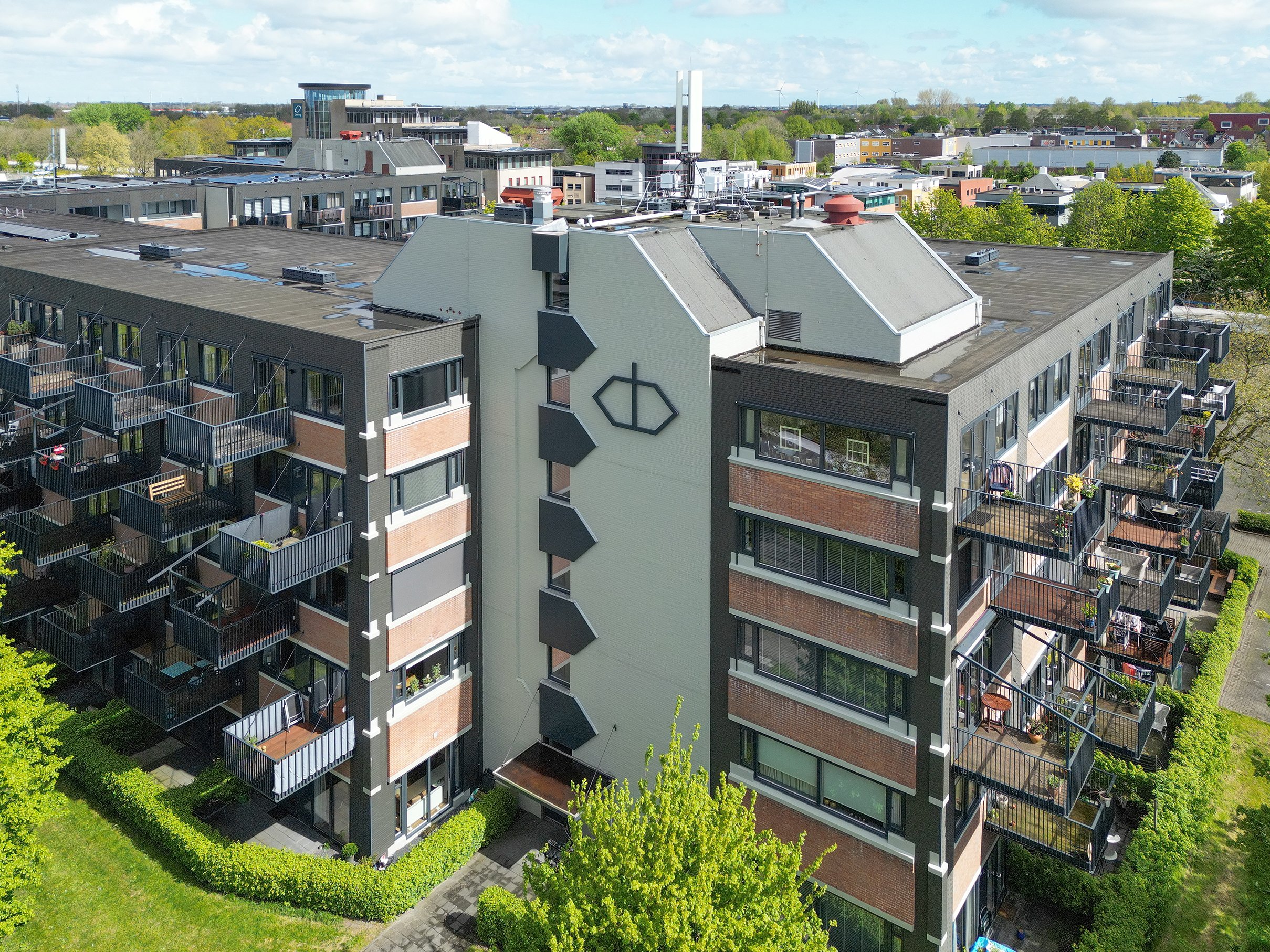 Luchtfoto van een moderne appartementengebouw met balkons en een tuin eronder, omgeven door groen en andere gebouwen in een frisse stadsomgeving onder een bewolkte hemel.