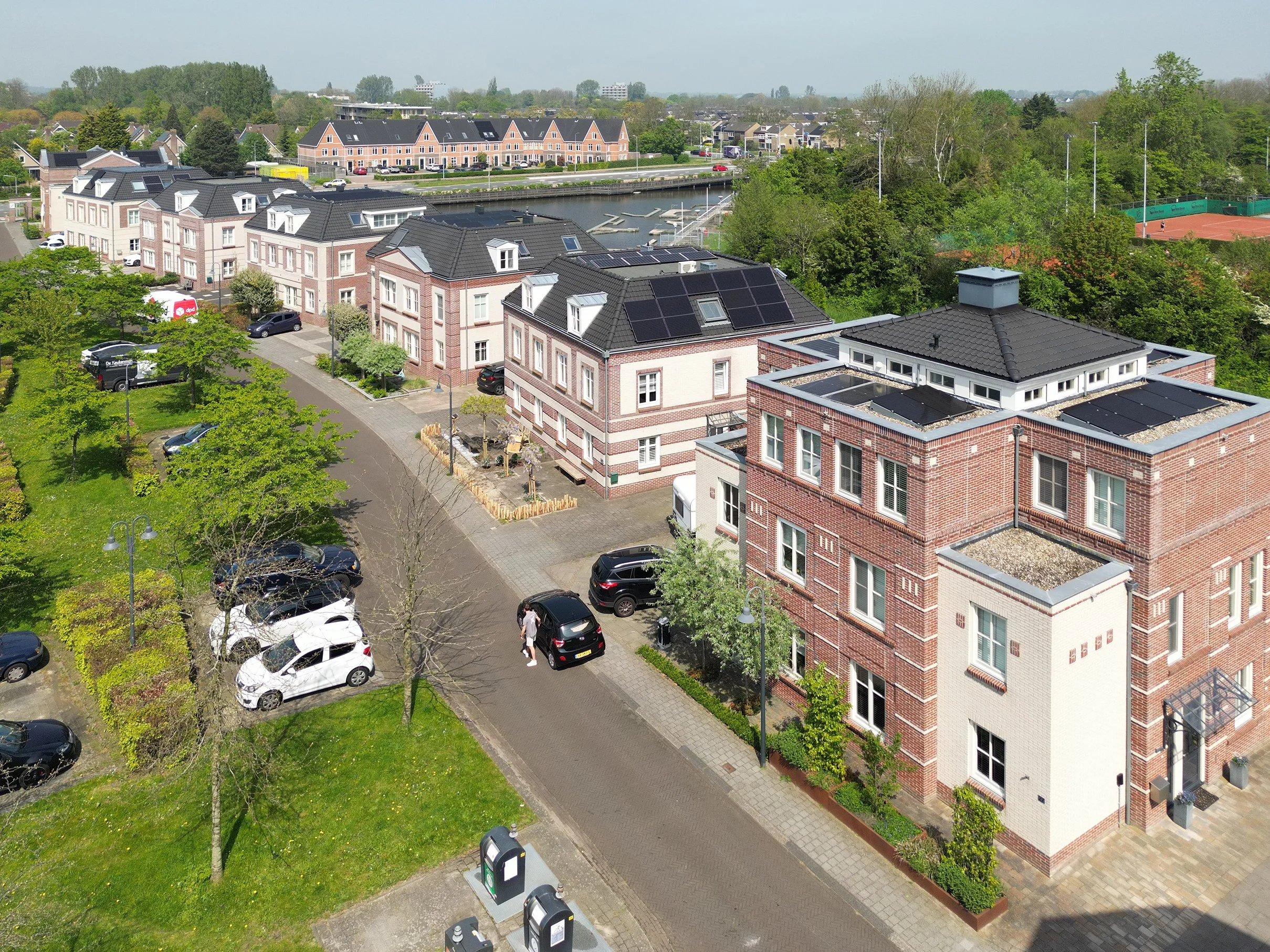 Luchtfoto van een woonwijk met rijtjeshuizen, een kanaal met boten, een tennisbaan en veel groen.