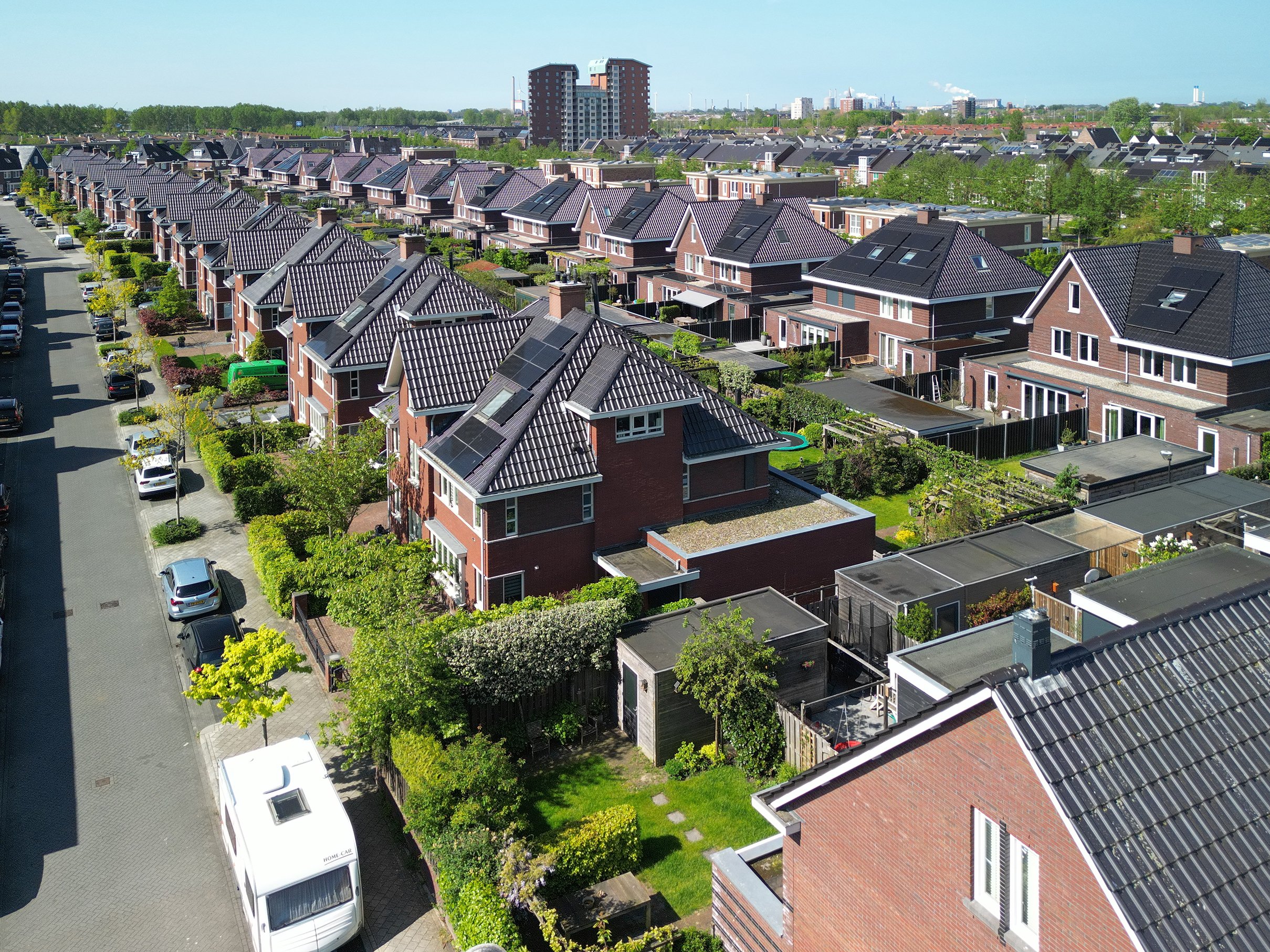 Luchtfoto van een woonwijk met rijtjeshuizen met daken met zonnepanelen, een parkeerplein met auto's, groene bomen en een stad op de achtergrond.
