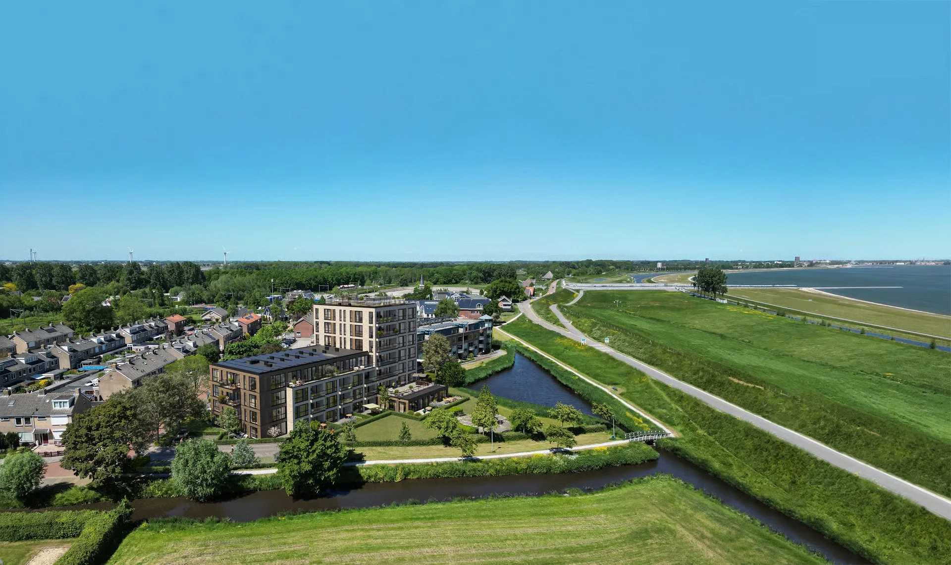 Luchtfoto van een dorp met moderne appartementen, groene gebieden, een rivier, een windmolenpark op de achtergrond, en een grote waterpartij aan de kust onder een heldere blauwe hemel.