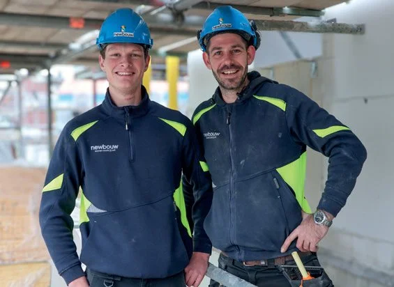 Twee mannen in bouwhelmen en werkpakken staan binnen een bouwplaats, lachend naar de camera.