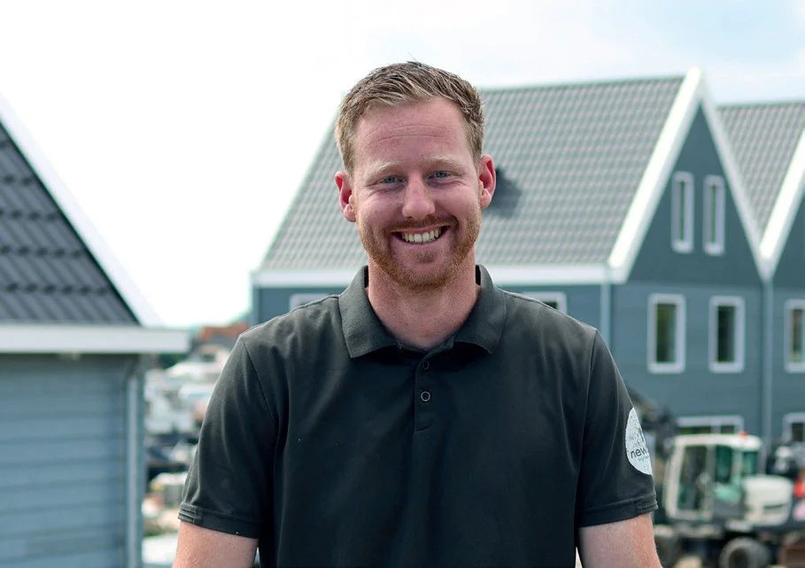 Man met rood haar en baard die glimlacht, staan voor rieten huizen op een zonnige dag.