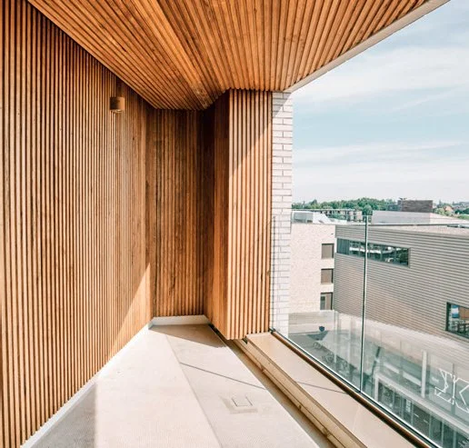 Een modern balkon met houten wand en plafond, afgezien van een glazen balustrade, met een uitzicht op gebouwen en een blauwe lucht.