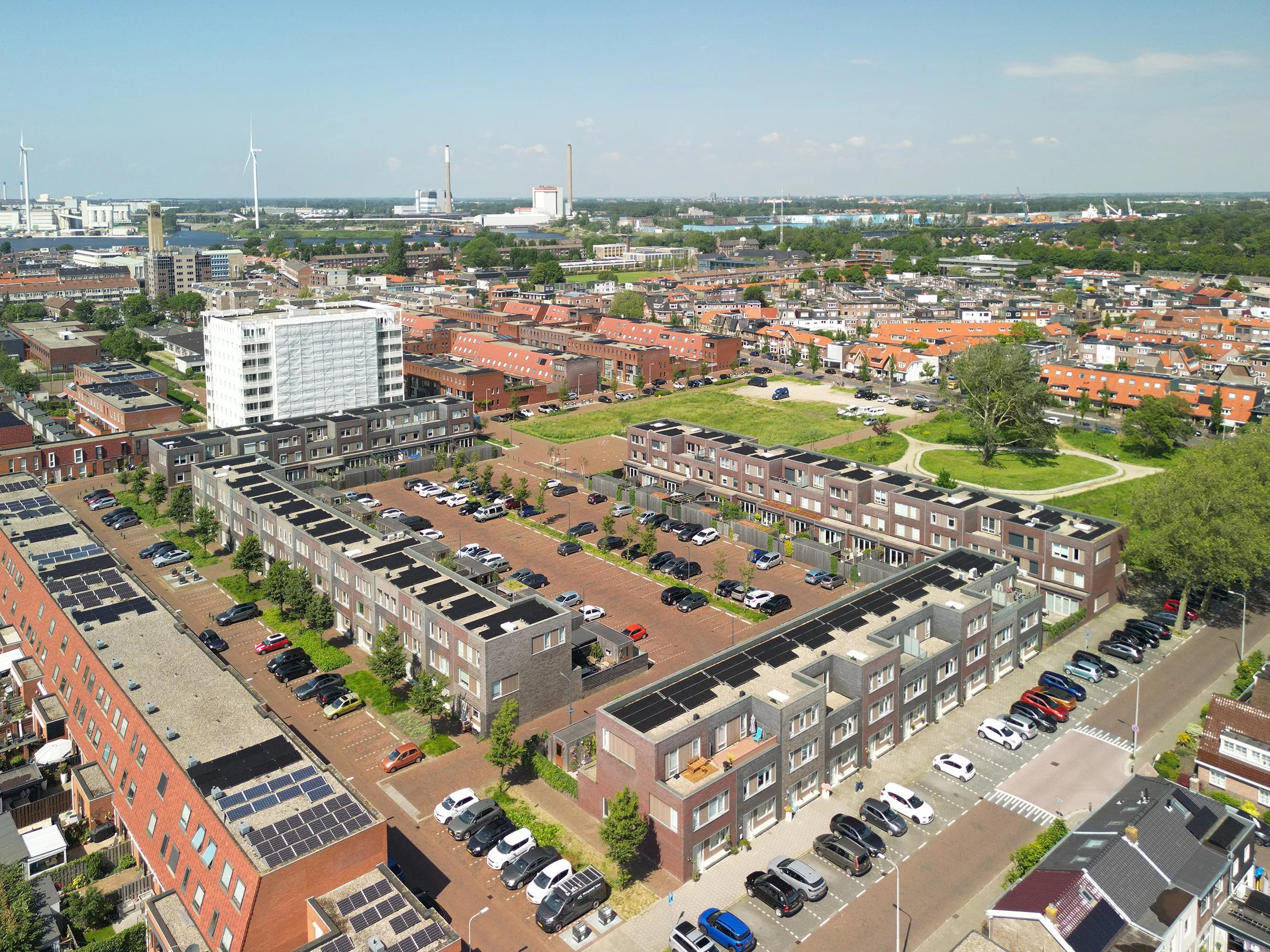 Uitzicht over een woonwijk met rijen huizen met plat dak en een aantal parkeerplaatsen, met in de achtergrond een industrieterrein met windmolens, fabrieken en een haven.