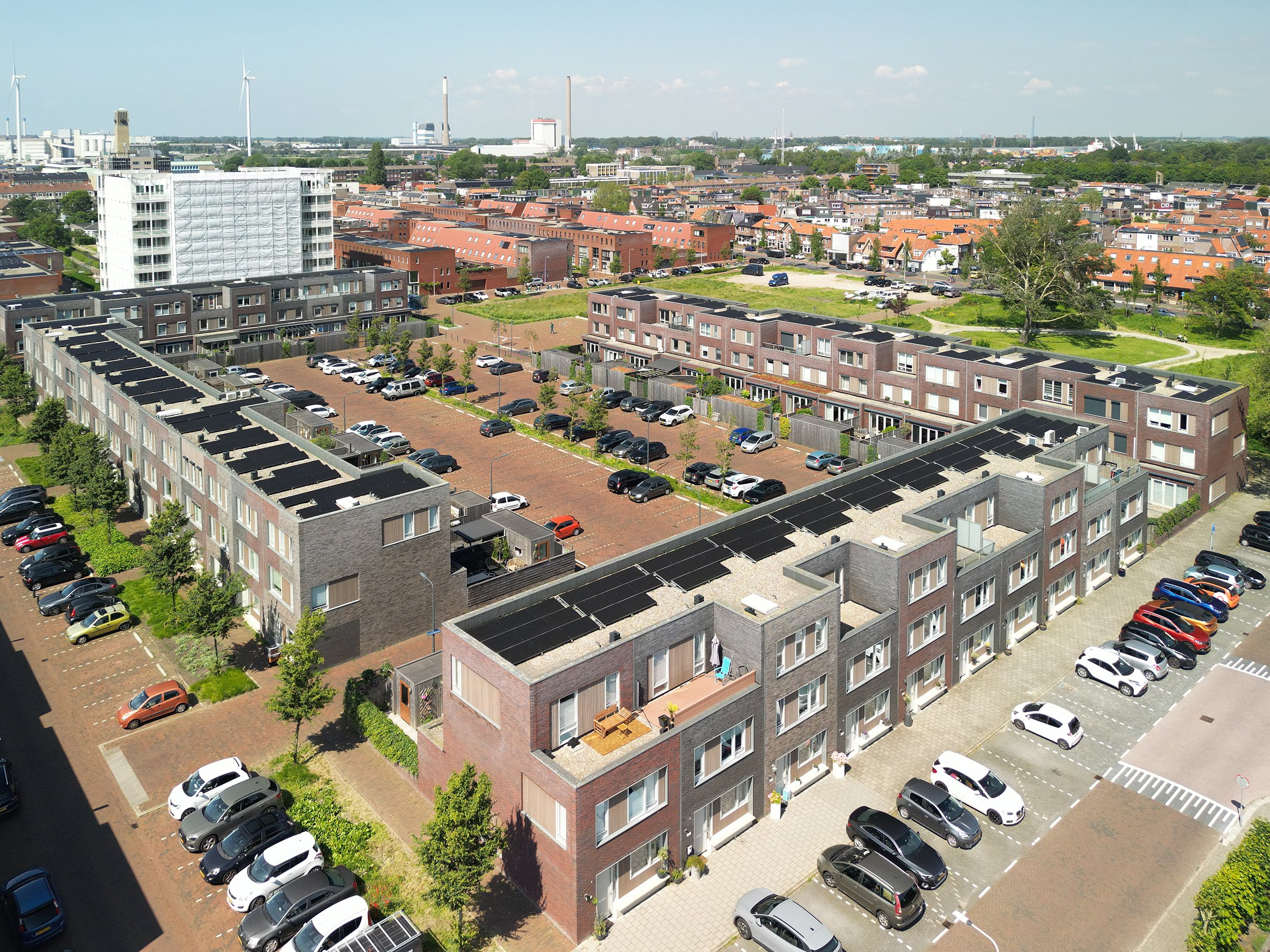 Luchtfoto van een woonwijk met meerdere flatgebouwen, parkeerplaatsen en een park met een grote boom. Op de achtergrond industriële gebouwen en windturbines.