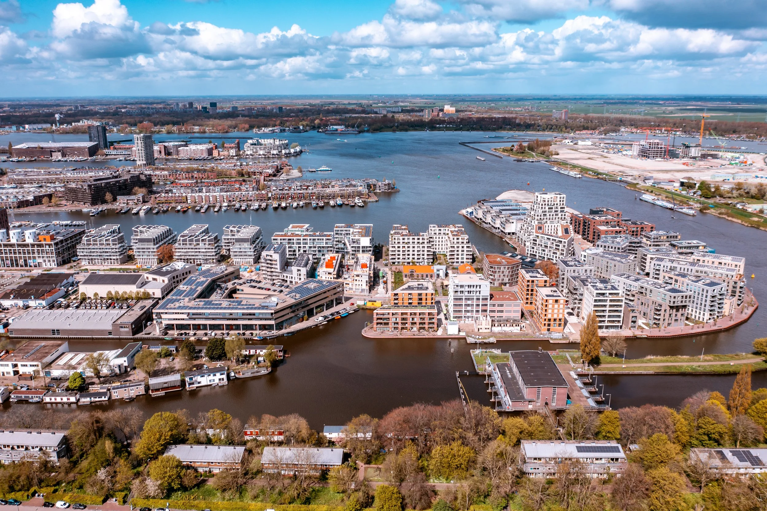 Luchtfoto van een stad met een rivier, moderne appartementen, boten, havens en een verder uitgestrekte landschappen met velden onder een deels bewolkte blauwe hemel.