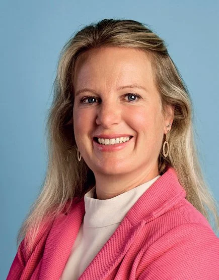 Portret van een glimlachende vrouw met lang blond haar, dragen een witte top en een roze jas, voor een lichtblauwe achtergrond.