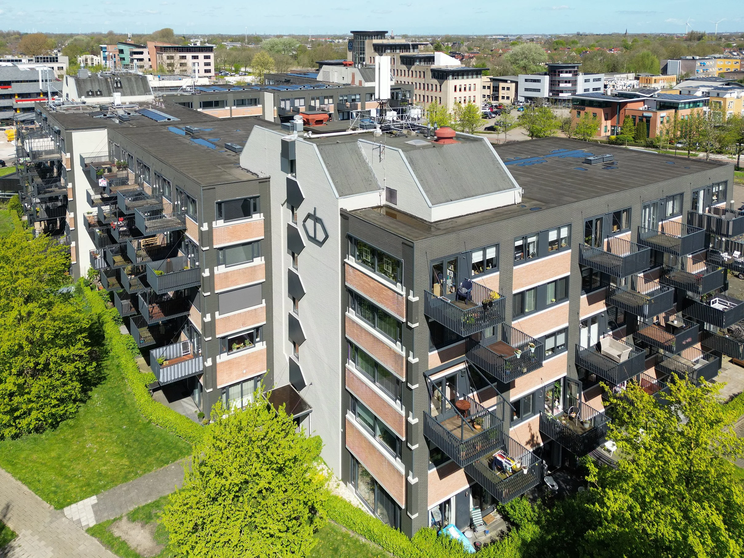 Hoge appartementencomplex met balkons, omgeven door groene bomen en een grasveld, in een stedelijk gebied met andere gebouwen op de achtergrond.