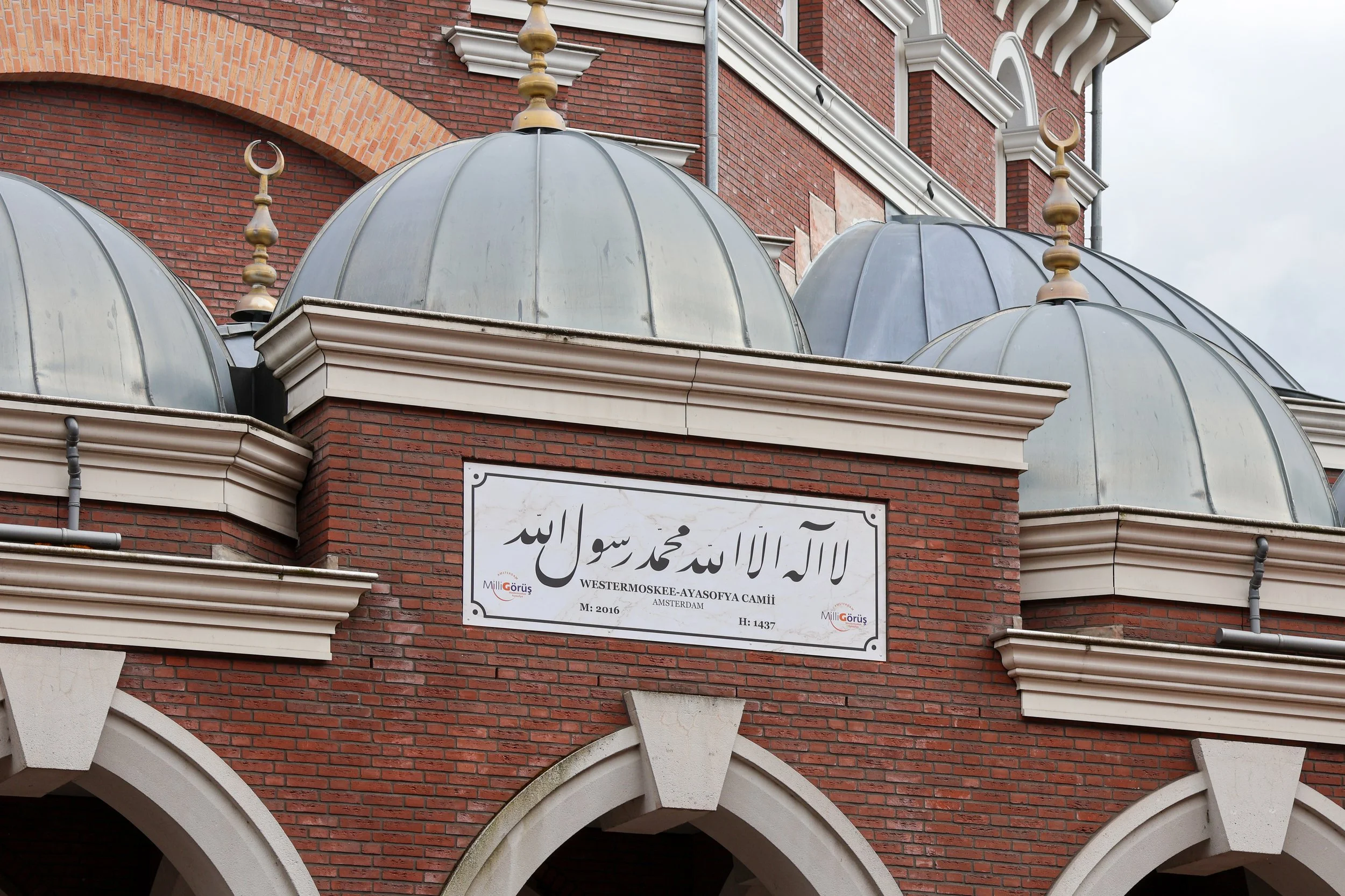 Uitzicht op een moskee met een gevel van rode bakstenen en drie grote metalen koepels met gouden ornamenten en halve maansymbolen. Op een bord staat Arabische tekst en informatie over de moskee, zoals naam, locatie en bouwjaar, in het Duits en Turks.
