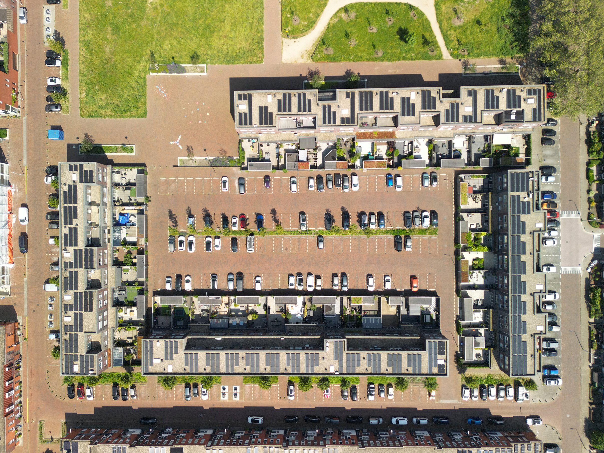 Een luchtfoto van een woonblok met een parkeerterrein, groene grasvelden, en omringende straten met geparkeerde auto's.