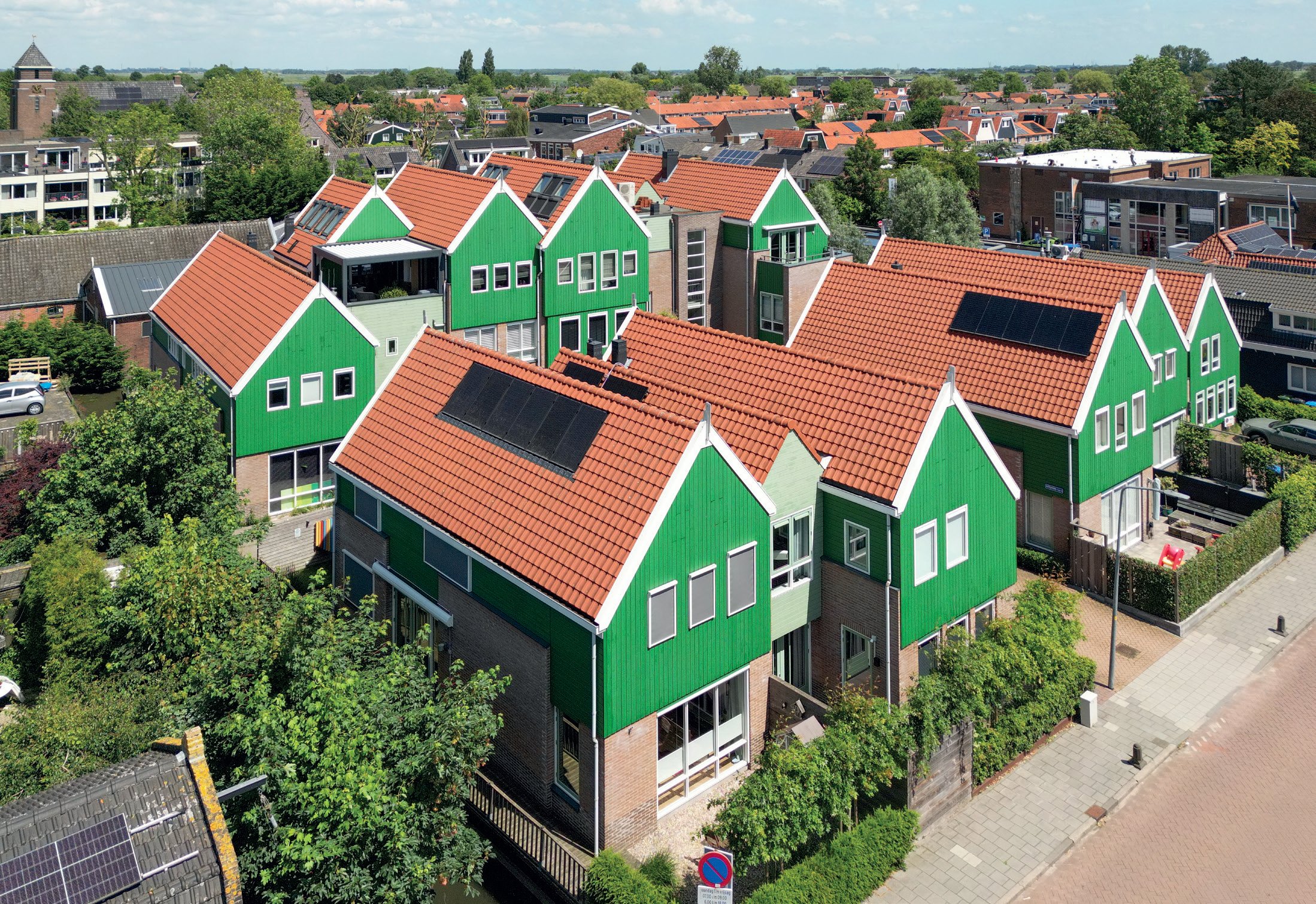 Uitzicht op een woonwijk met groene huizen met rode daken, omringd door bomen en overige huizen met zonnepannen op het dak, onder een bewolkte blauwe hemel.