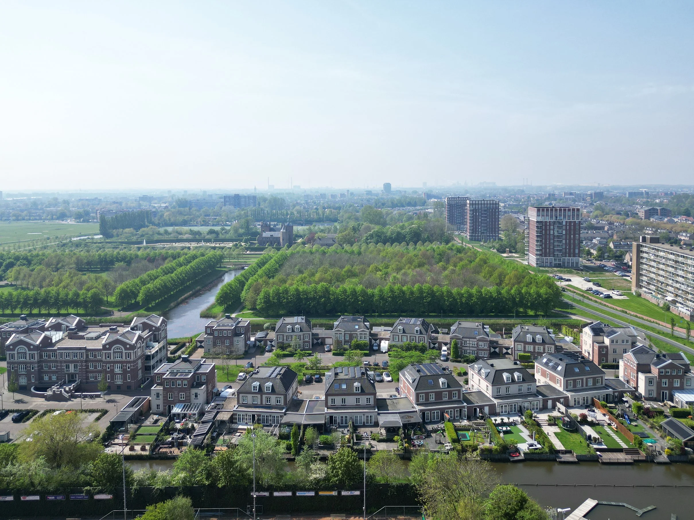 Uitzicht op een stad met woongebouwen, een rivier, groene parken en bomen onder een lichte blauwe hemel.