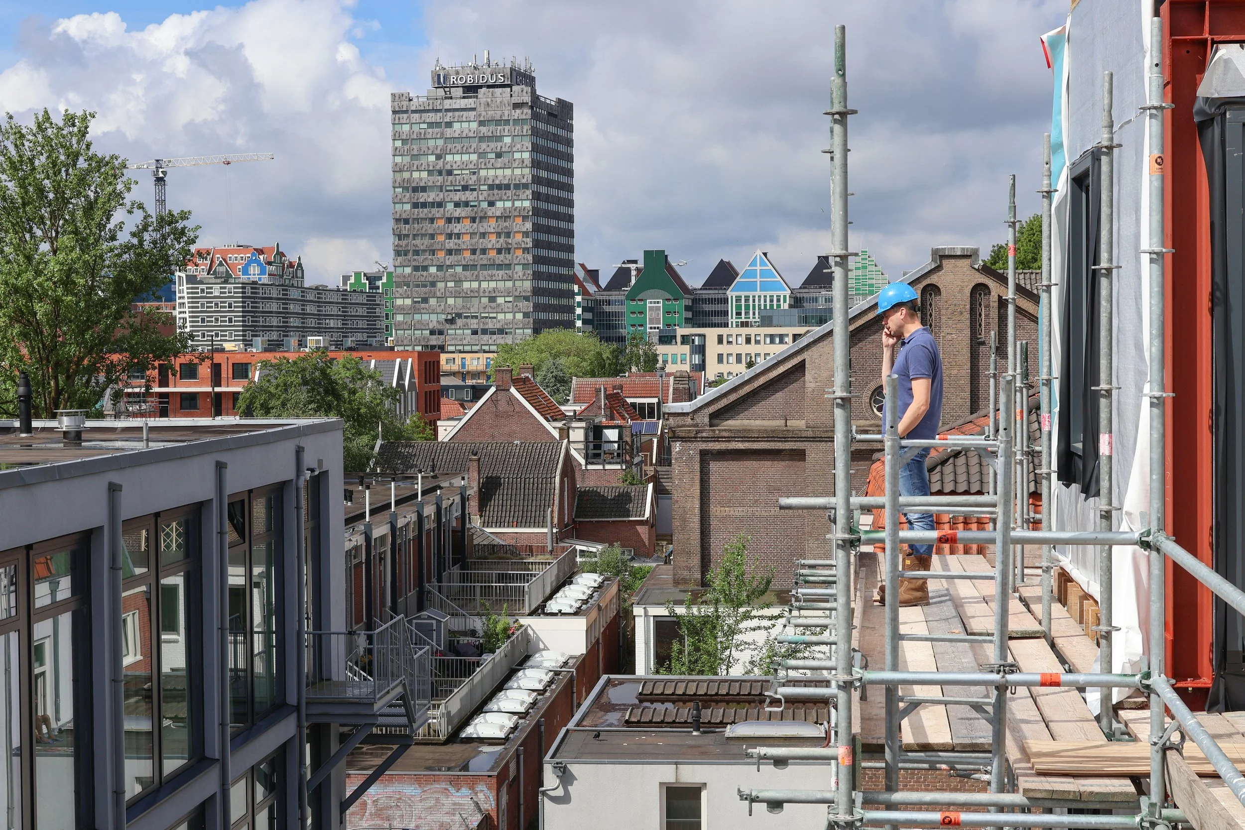 Man met blauwe helm praat op mobiel op dak met steigers, uitzicht op stad met hoge en lage gebouwen en wolken in de lucht.