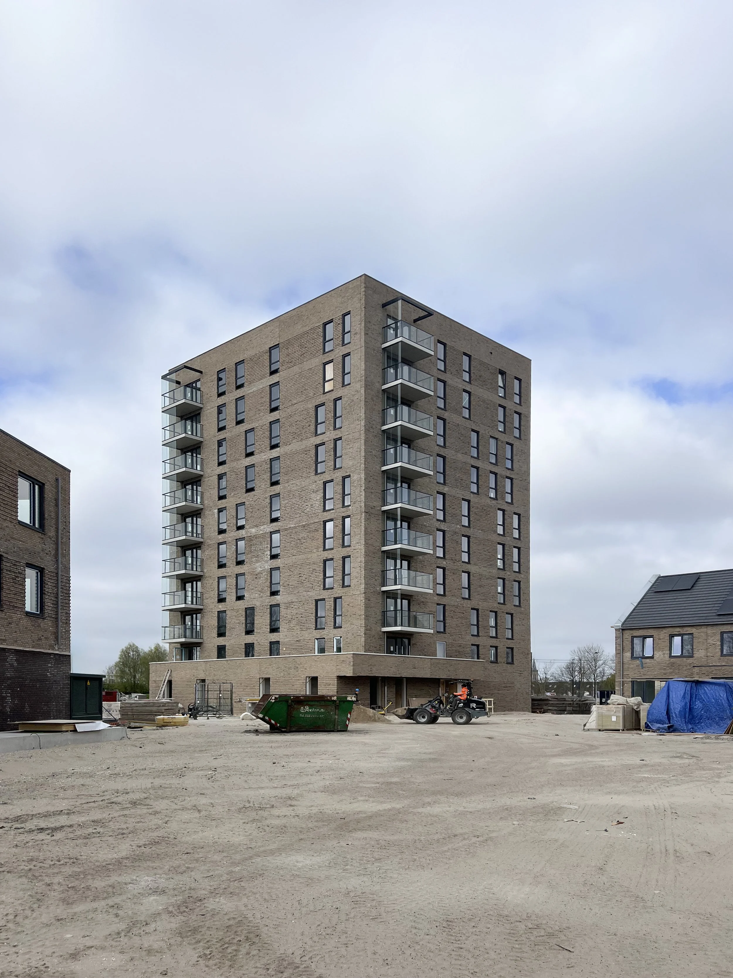 Een hoog gebouw onder constructie in een bouwplaats met bouwmaterialen, een container, en bouwmachines op de voorgrond.