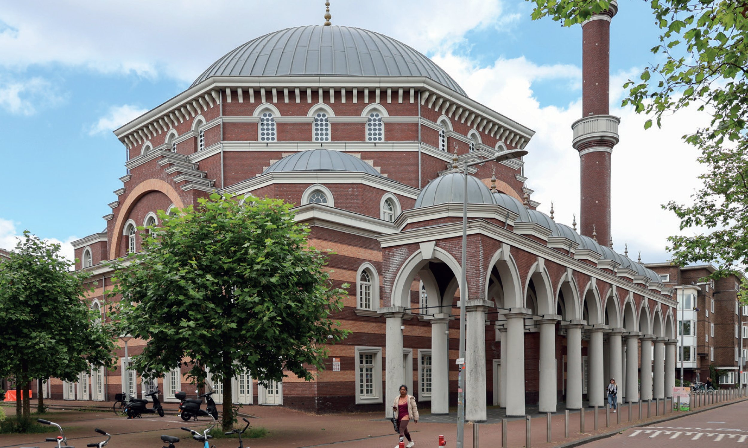 Een grote moskee met een ronde koepel en een minaret, omringd door bomen, met mensen en fietsen op de voorgrond, onder een partly bewolkte blauwe hemel.