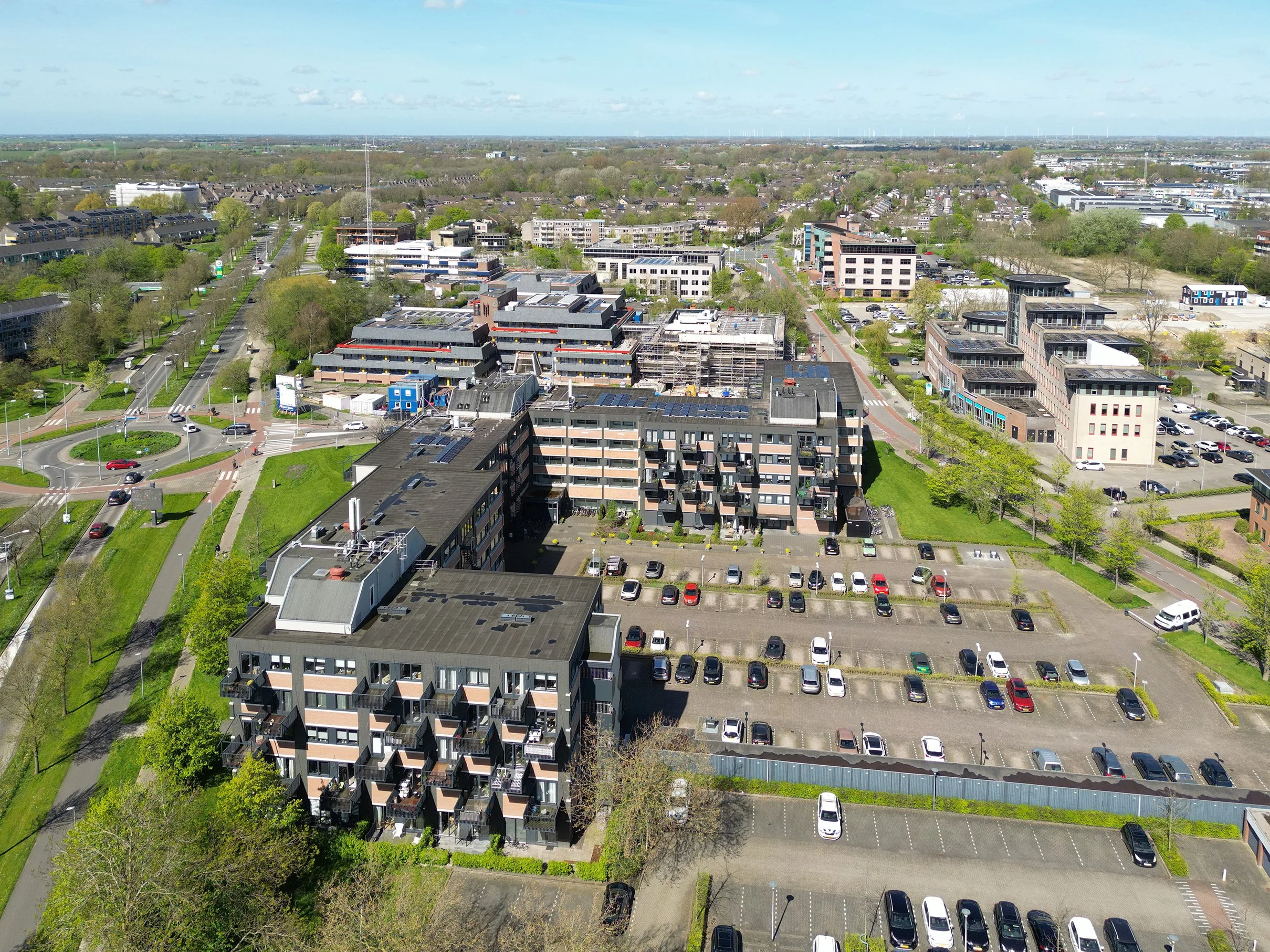 Luchtfoto van een stad met verschillende gebouwen, wegen en groene bomen onder een gedeeltelijk bewolkte blauwe lucht.