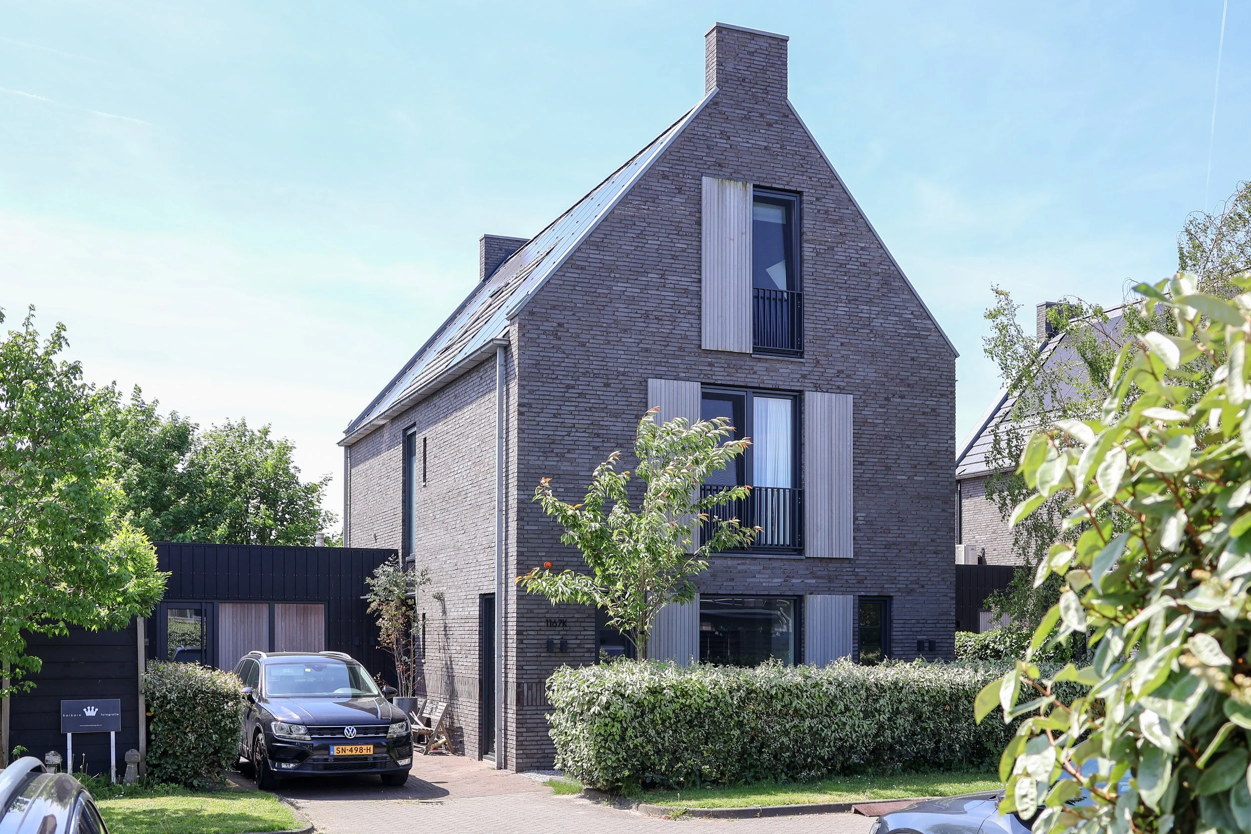 Moderne woning met een gevel van baksteen en grote ramen, omgeven door bomen en een voortuin, met geparkeerde auto's op de oprit.
