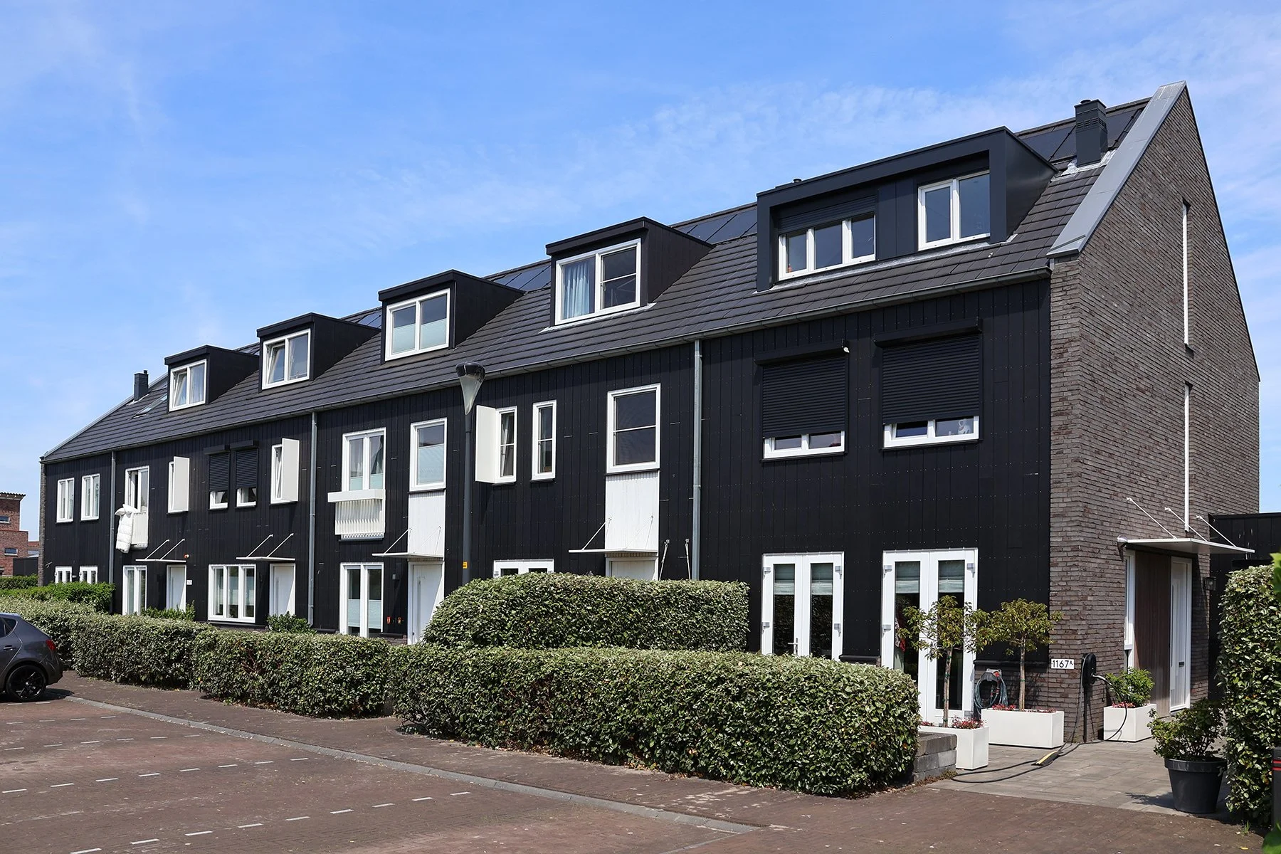 Rij van moderne zwarte appartementen met witte ramen, met een voorhof met stoep, struiken, een geparkeerde zwarte auto, en een blauwe hemel.