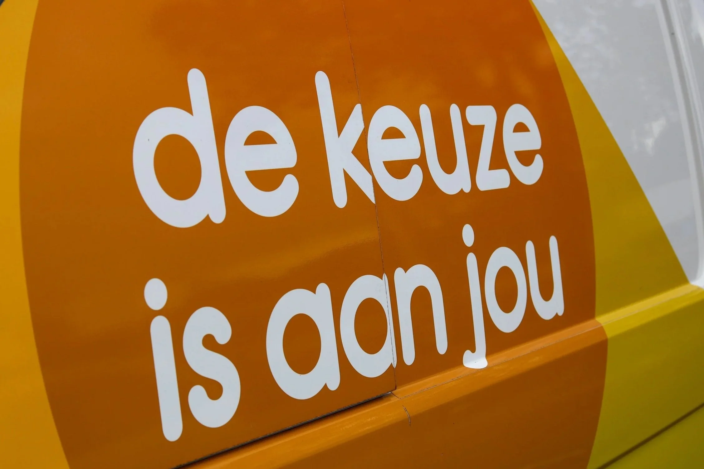 Afbeelding van een oranje bord met de woorden 'de keuze' in wit, gedeeltelijk zichtbaar.
