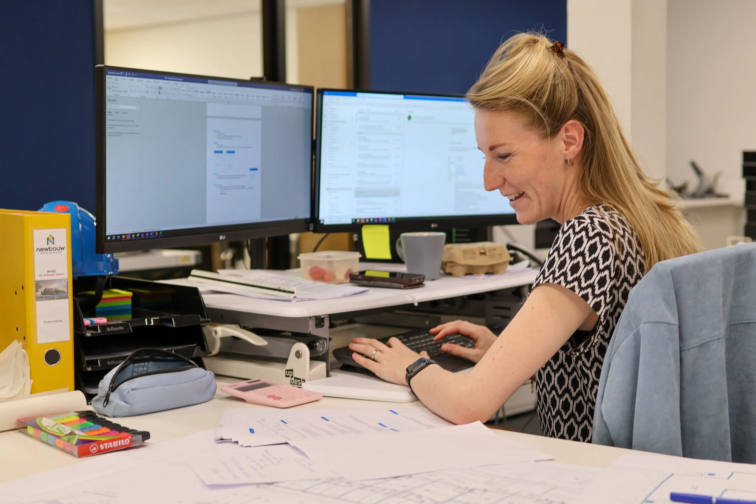 Een vrouw zit aan een bureau met twee computermonitoren, aan het schrijven op haar toetsenbord. Op het bureau liggen papieren, een beker, een notitieboekje, opladers en schrijfbenodigdheden.