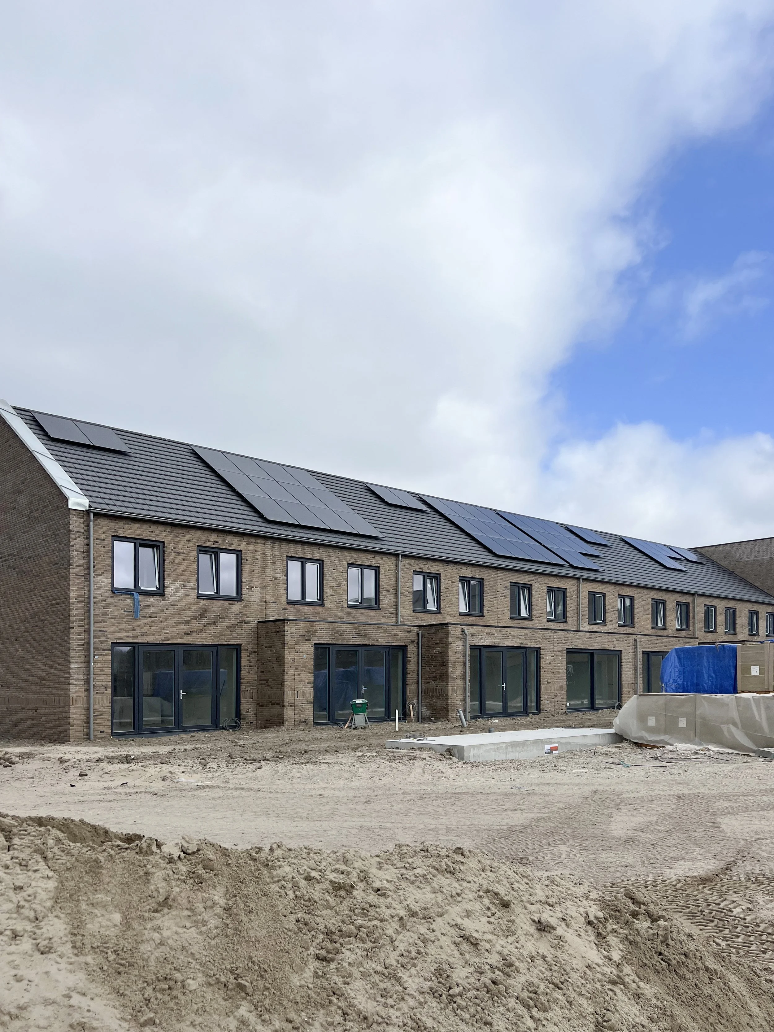 Nieuw gebouw onder constructie met zonnepanelen op het dak, bouwmateriaal en bouwmaterialen op de voorgrond, en een deels bewolkte hemel erboven.