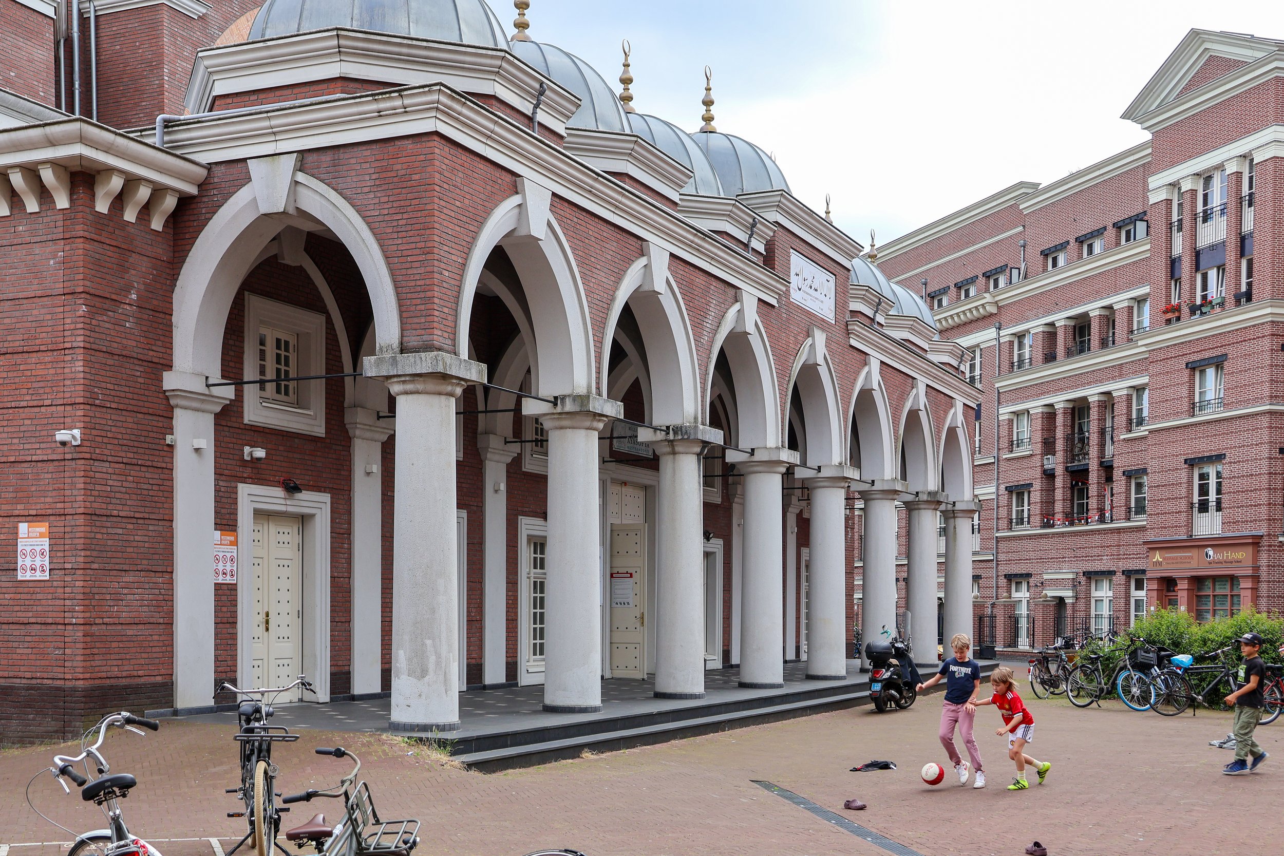 Jeugd speelt voetbal op straat voor een gebouw met bekende architectuur, omringd door fietsen en appartementen.