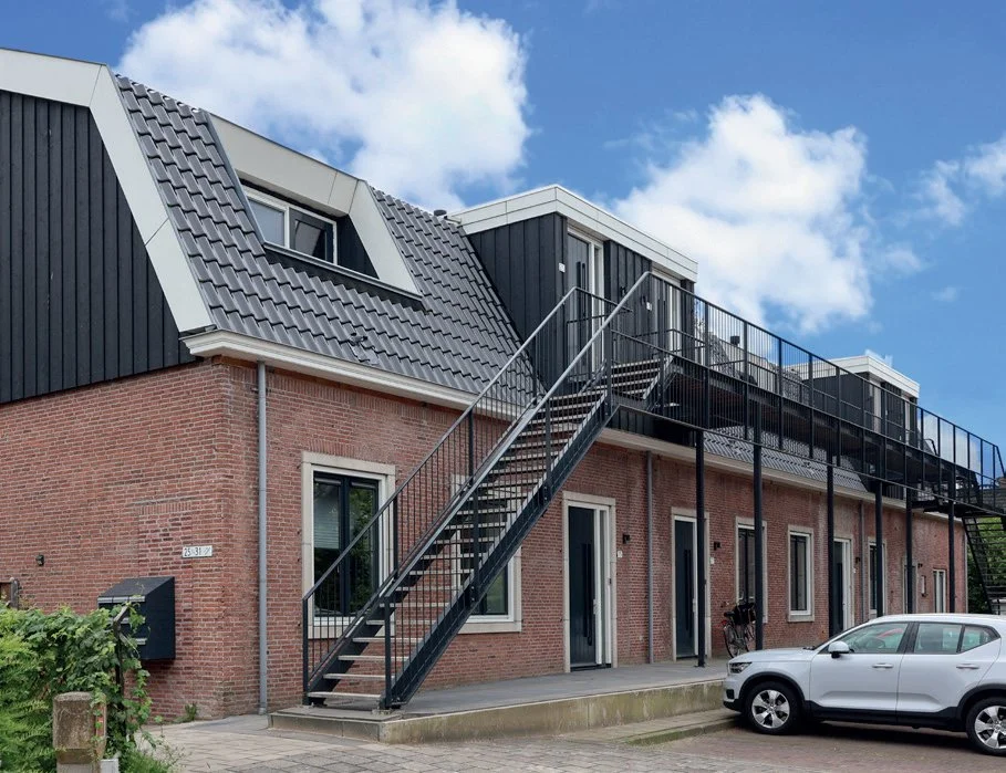 Een tweeverdienhuis met een randdak en een aparte bovenverdieping met een balkon, zwarte metalen trappen naar het balkon, een witte auto geparkeerd voor het huis.