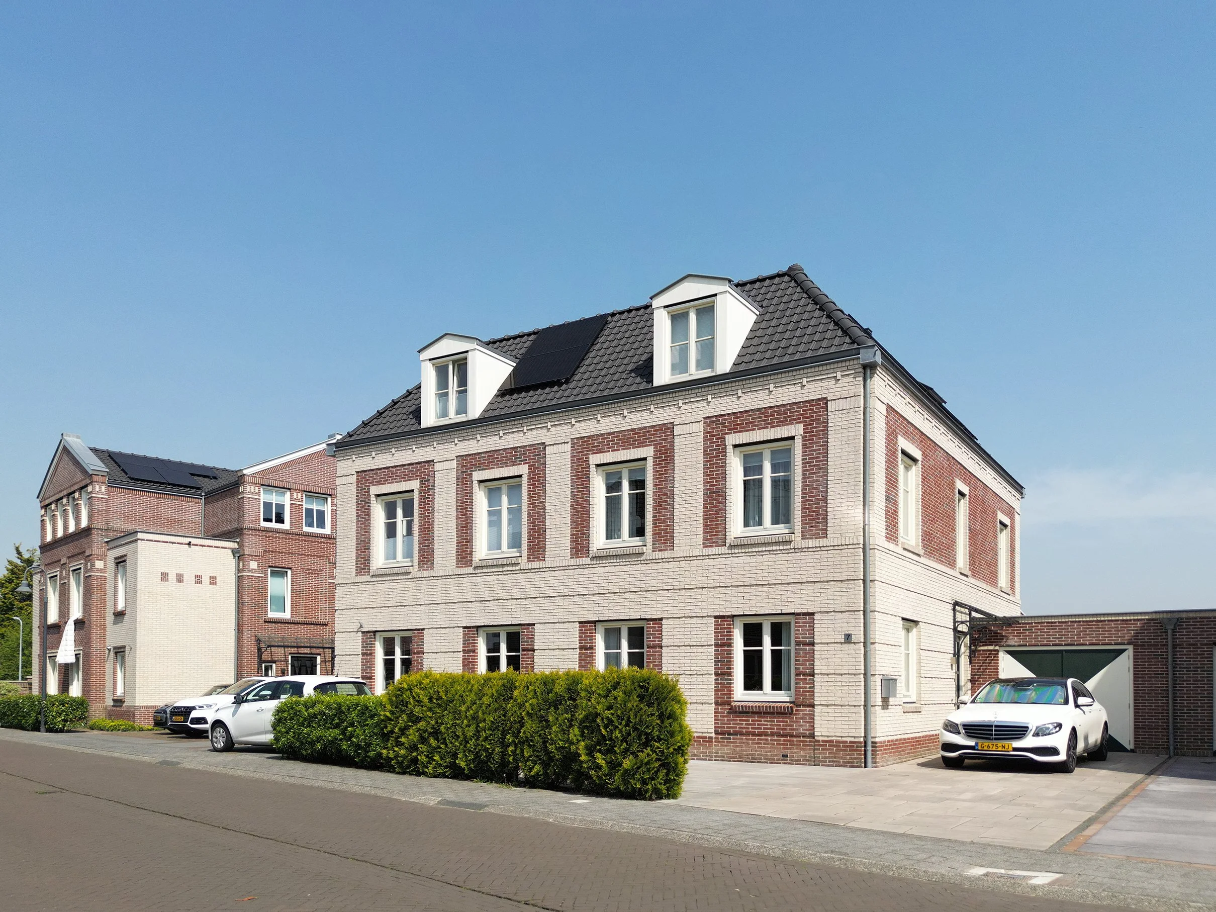Een wit en rood bakstenen gebouw met een zadeldak en een kleine dakraam, er staan enkele auto’s geparkeerd op de parkeerplaats voor het gebouw, er is een heg en een laag groen struikje voor het gebouw, en de lucht is helder en blauw.